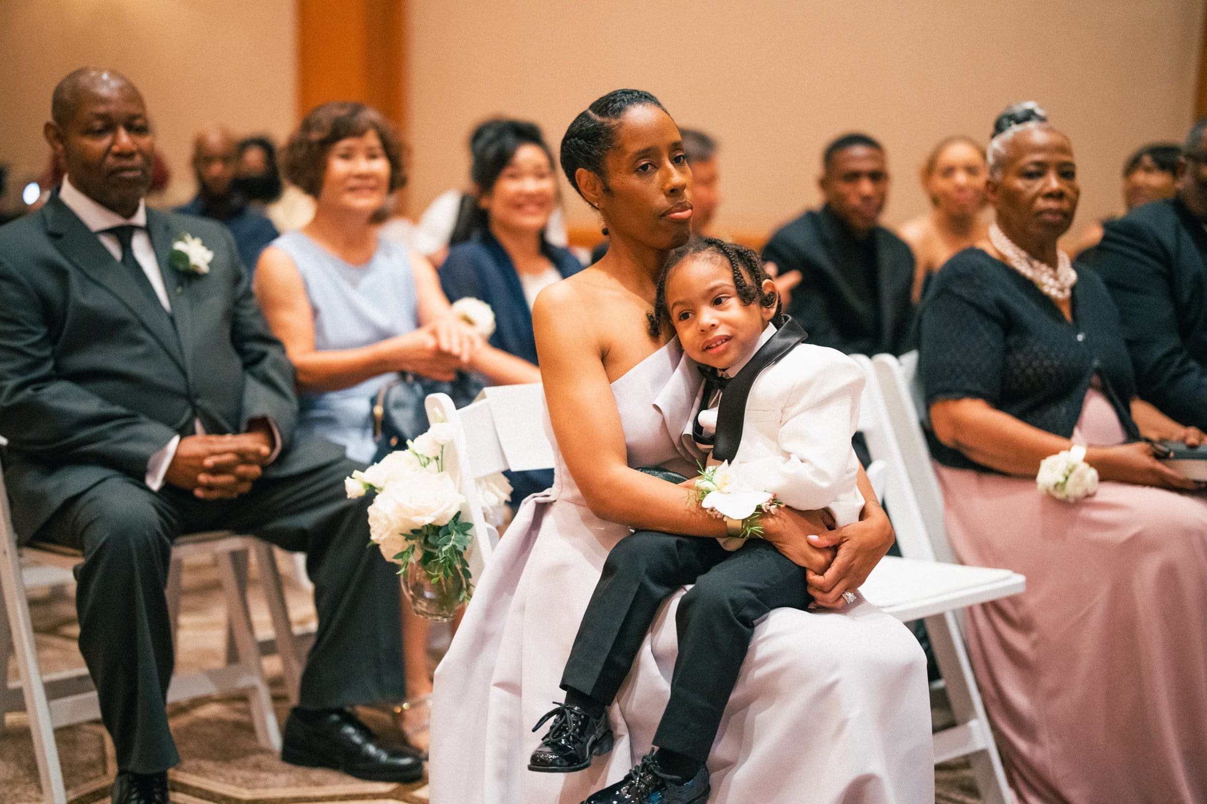 umstead wedding photography - kayla & deandre At the Umstead wedding, a woman seated in a formal setting holds a young boy on her lap. Both are elegantly dressed, with the boy in a white jacket. They are surrounded by other seated guests, all attired for this special occasion.