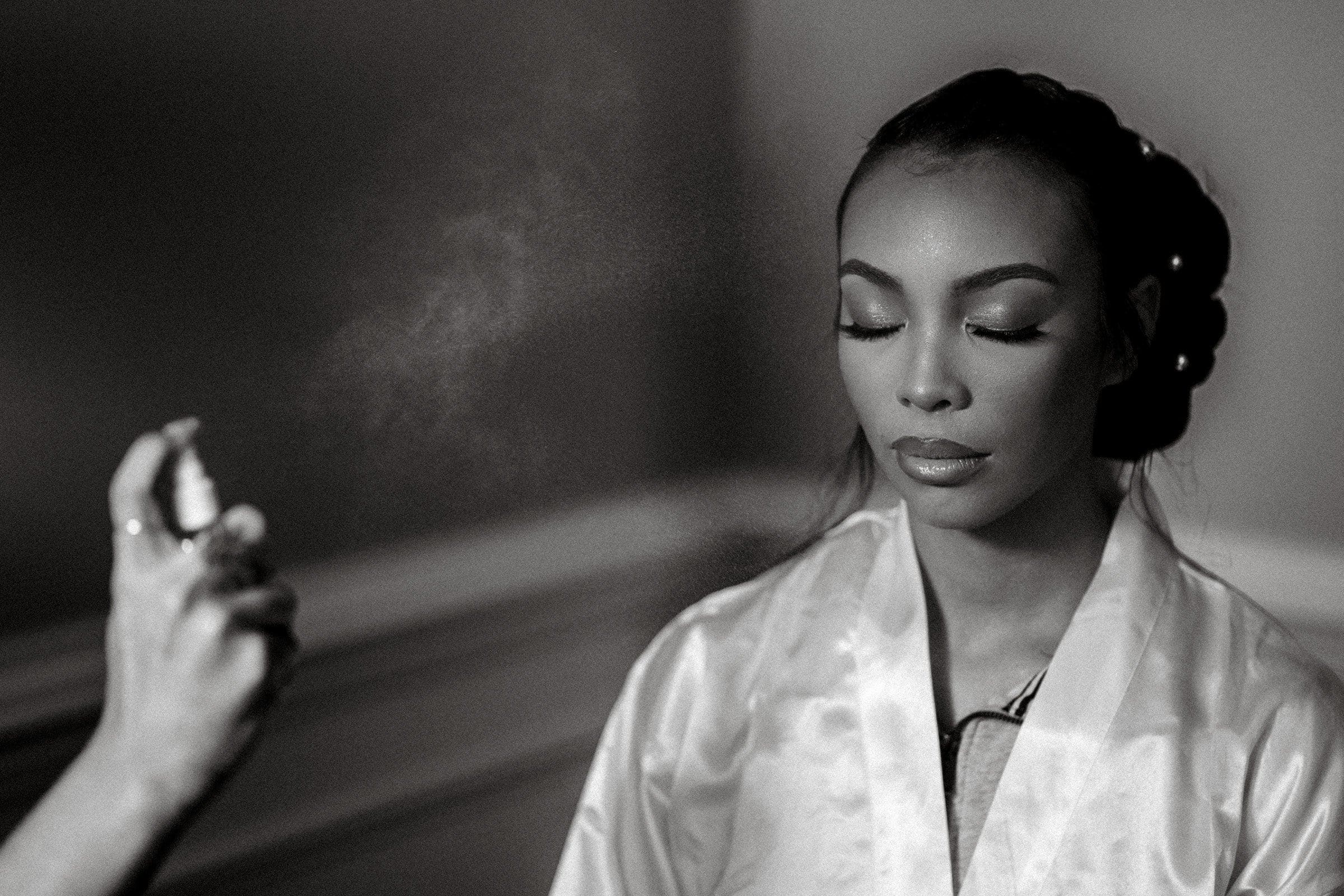 umstead wedding photography - kayla & deandre A woman in a satin robe with her eyes closed is getting her makeup set with a spray, her hair elegantly styled. The scene exudes the calm and focused atmosphere of an Umstead wedding preparation. Grayscale image.