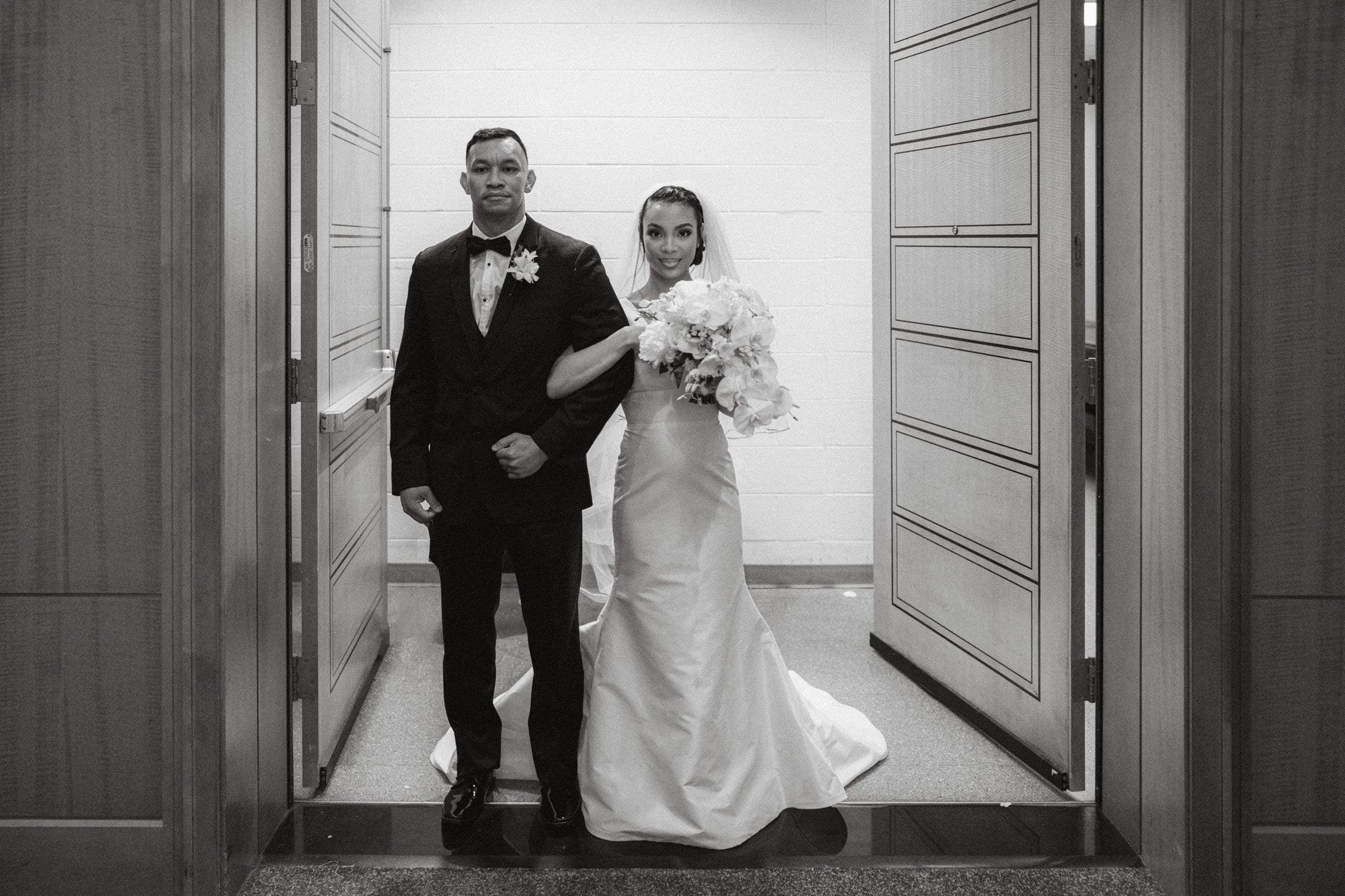 umstead wedding photography - kayla & deandre A bride and groom stand together in a doorway during their Umstead wedding. The bride wears a flowing white gown and holds a bouquet of flowers, while the groom is dressed in a black tuxedo. Both look forward, appearing composed and elegant.