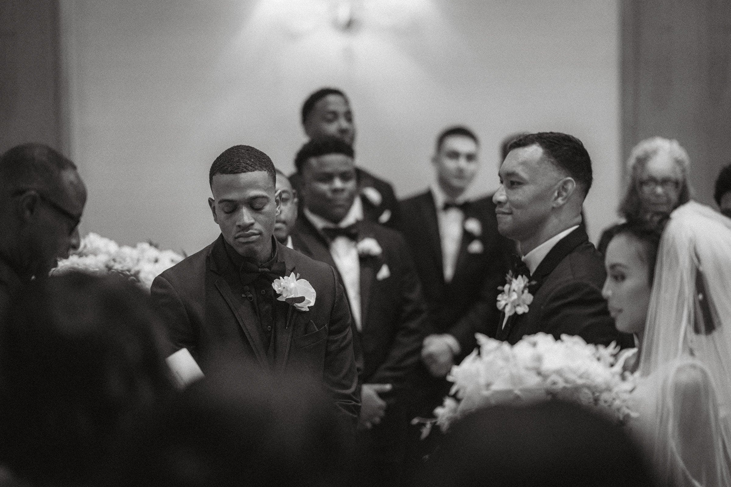 umstead wedding photography - kayla & deandre A black and white photo of an Umstead wedding ceremony captures a group of people, mostly in suits, surrounding the bride and groom. The groom smiles slightly while the bride looks down, all set against the backdrop of elegant floral arrangements.