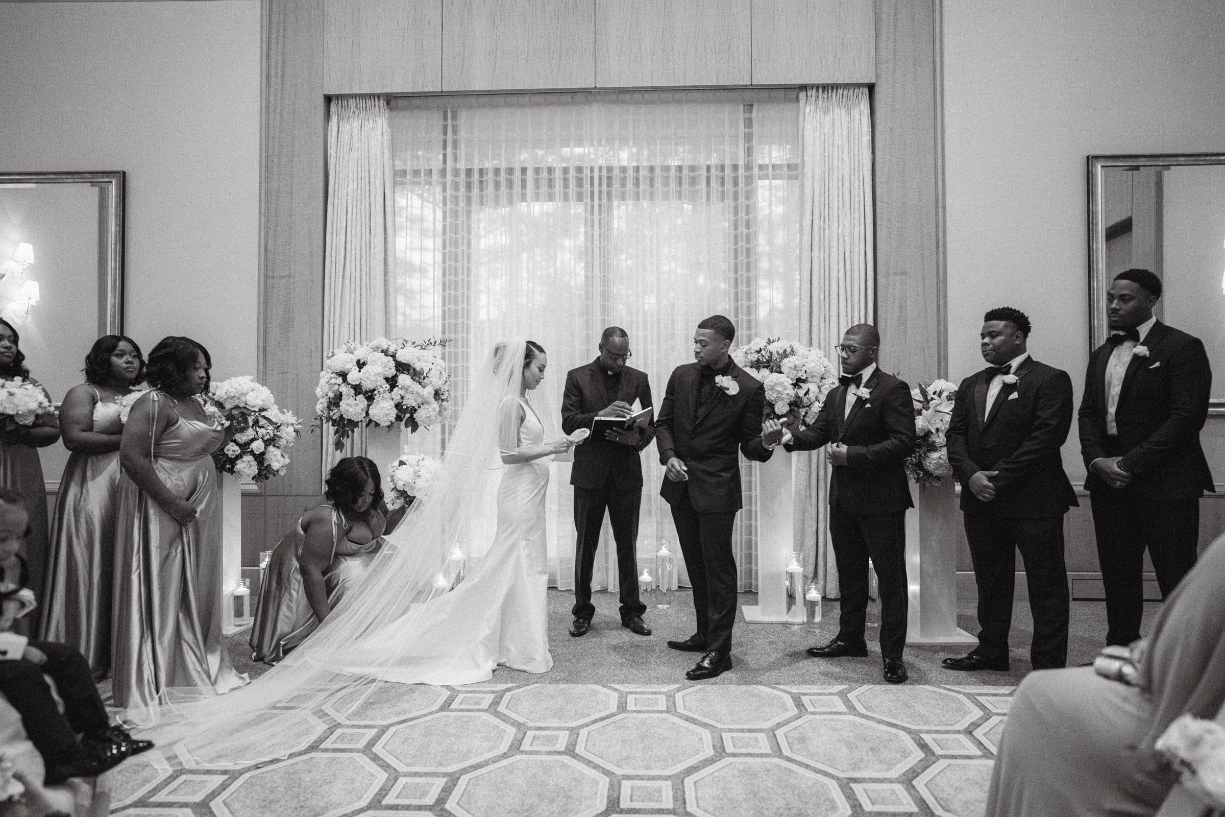 umstead wedding photography - kayla & deandre At an enchanting Umstead wedding, a bride and groom stand at the altar. The bride holds a microphone, while the groom gestures warmly. Bridesmaids and groomsmen surround them, framed by elegant floral arrangements and flickering candles in an intimate setting.