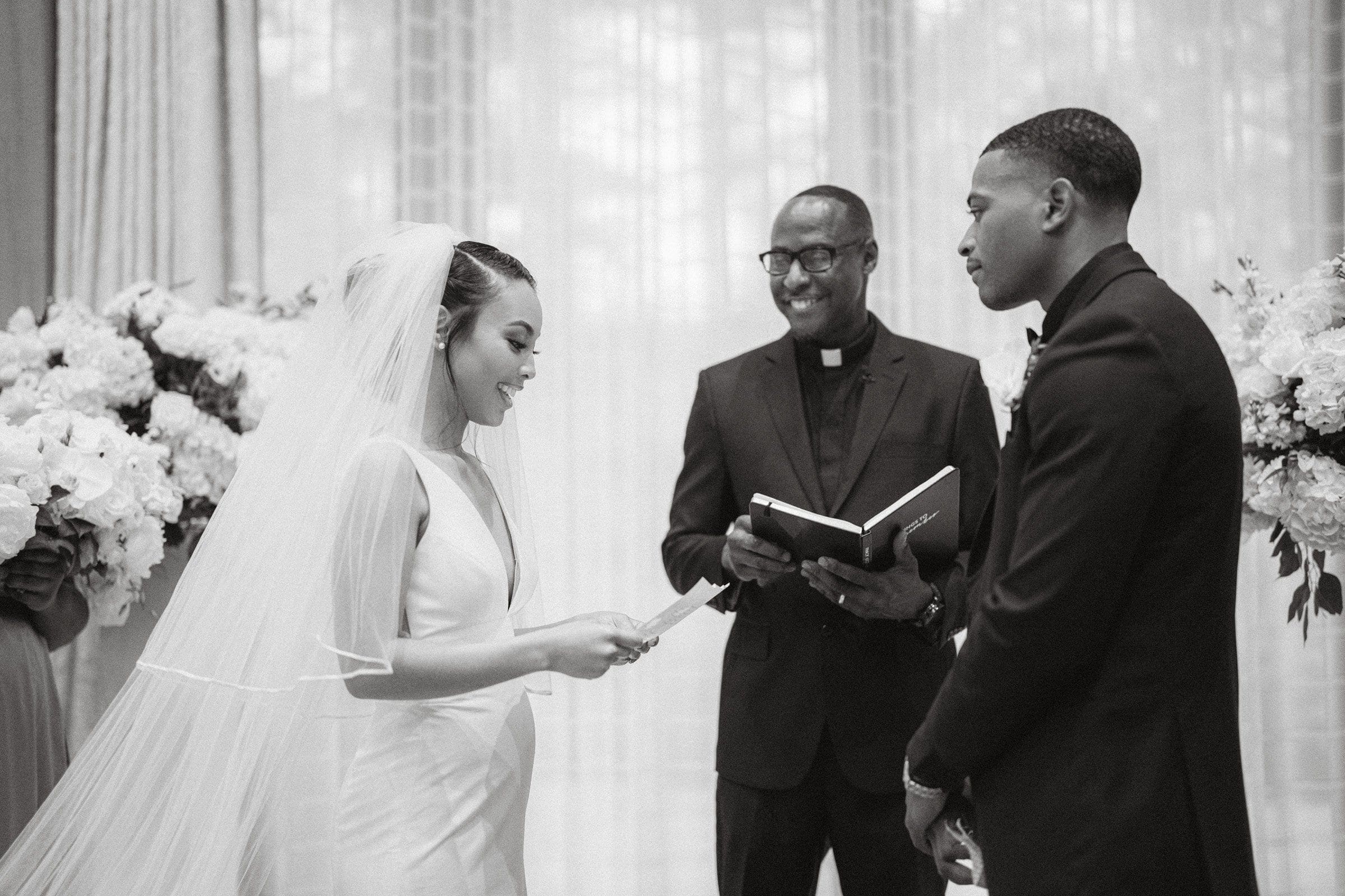 umstead wedding photography - kayla & deandre A bride and groom stand facing each other, smiling during their vows at an Umstead wedding. The pastor, holding a book, stands between them amidst elegant floral arrangements in the background.