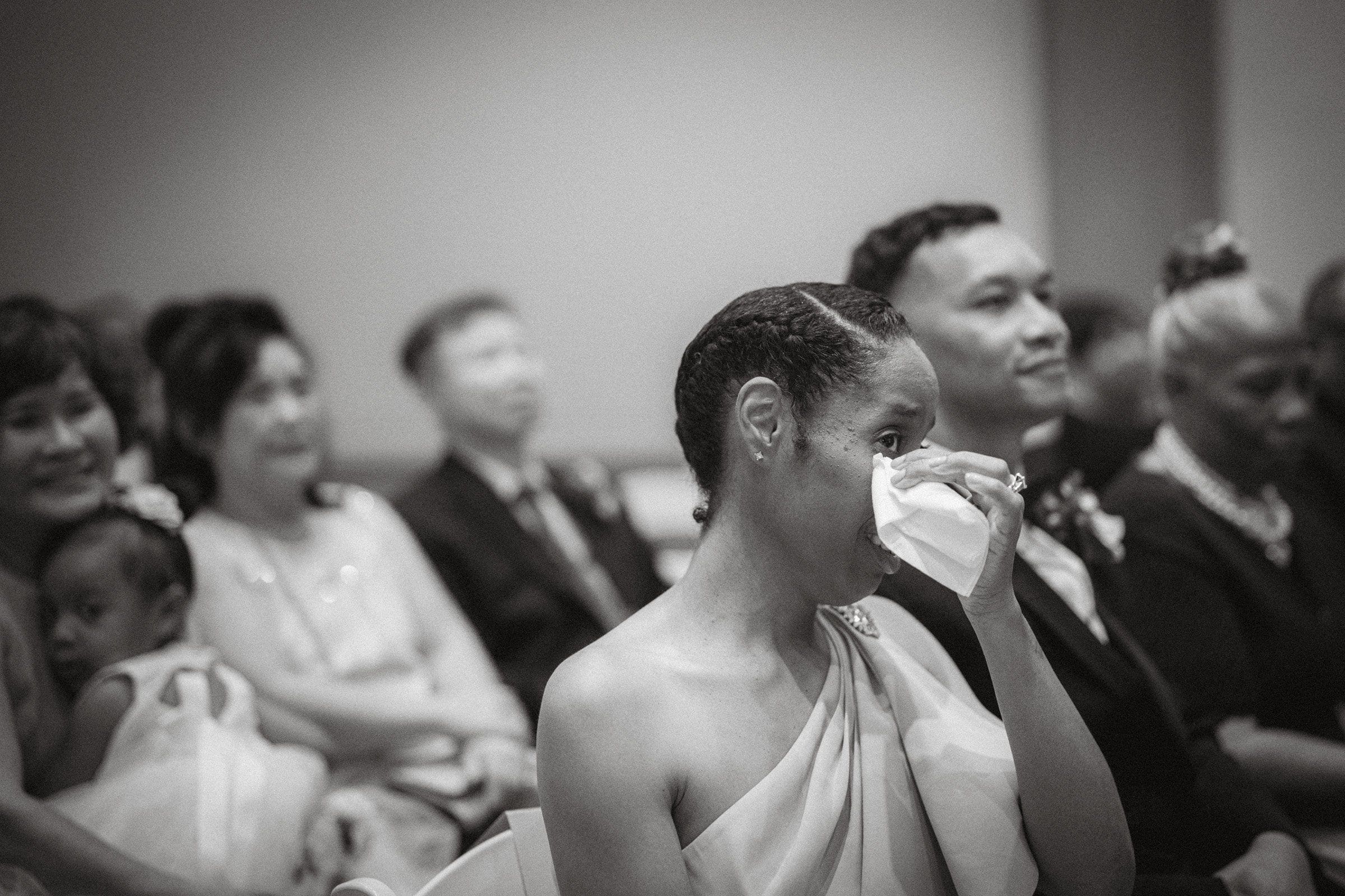umstead wedding photography - kayla & deandre A black and white photo captures the poignant moment of a woman in a one-shoulder dress wiping tears with a tissue during an Umstead wedding. She sits among other attendees, including a young child and several formally dressed adults, all united in the emotion of the event.