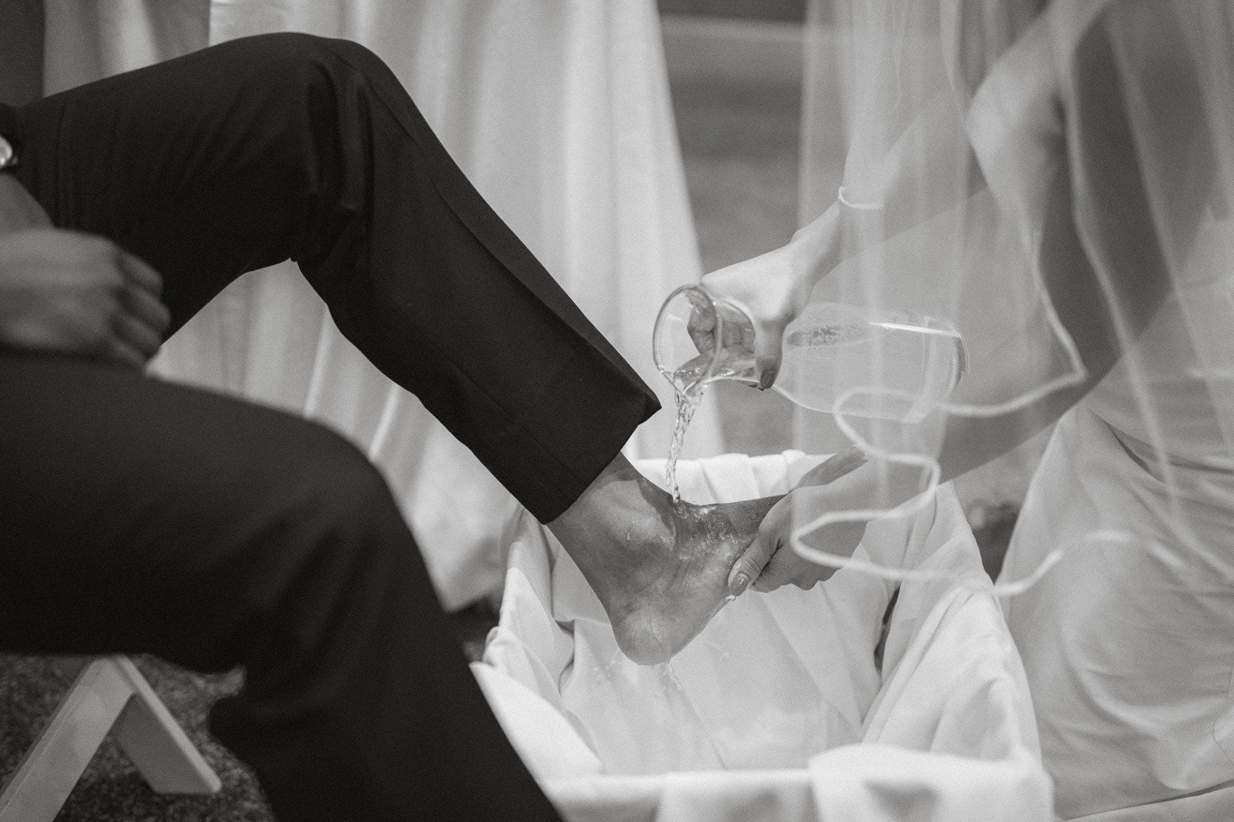 umstead wedding photography - kayla & deandre A black and white photo from an Umstead wedding captures a person pouring water from a jug over another's foot, resting in a basin lined with fabric. The seated individual wears dark pants, while the one performing this intimate ritual is partially veiled.