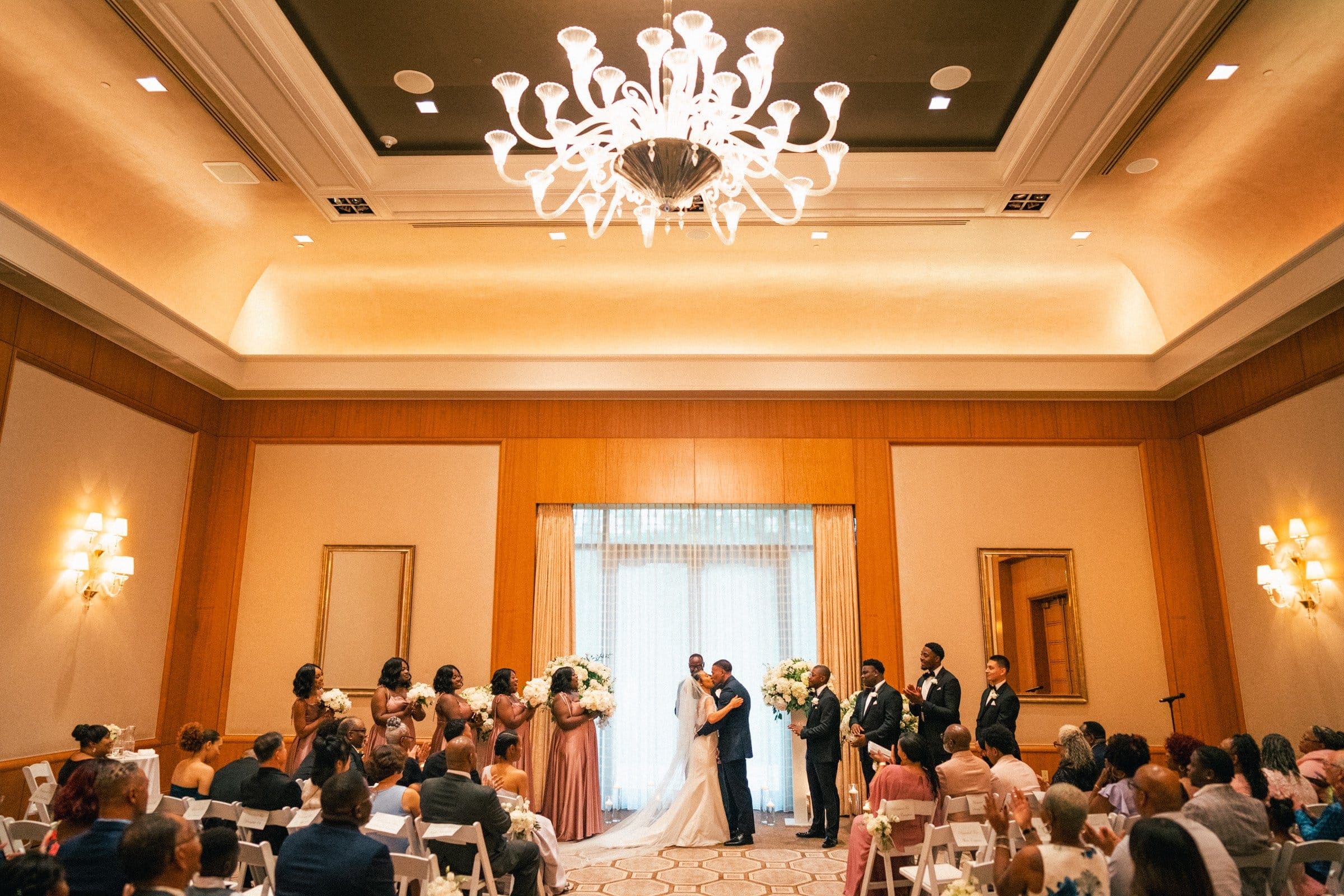 umstead wedding photography - kayla & deandre During their Umstead wedding, a couple kisses at the altar in a large, elegant room with a chandelier. Guests are seated, and the bridal party stands nearby. Bridesmaids in pink dresses and groomsmen in gray suits look on.