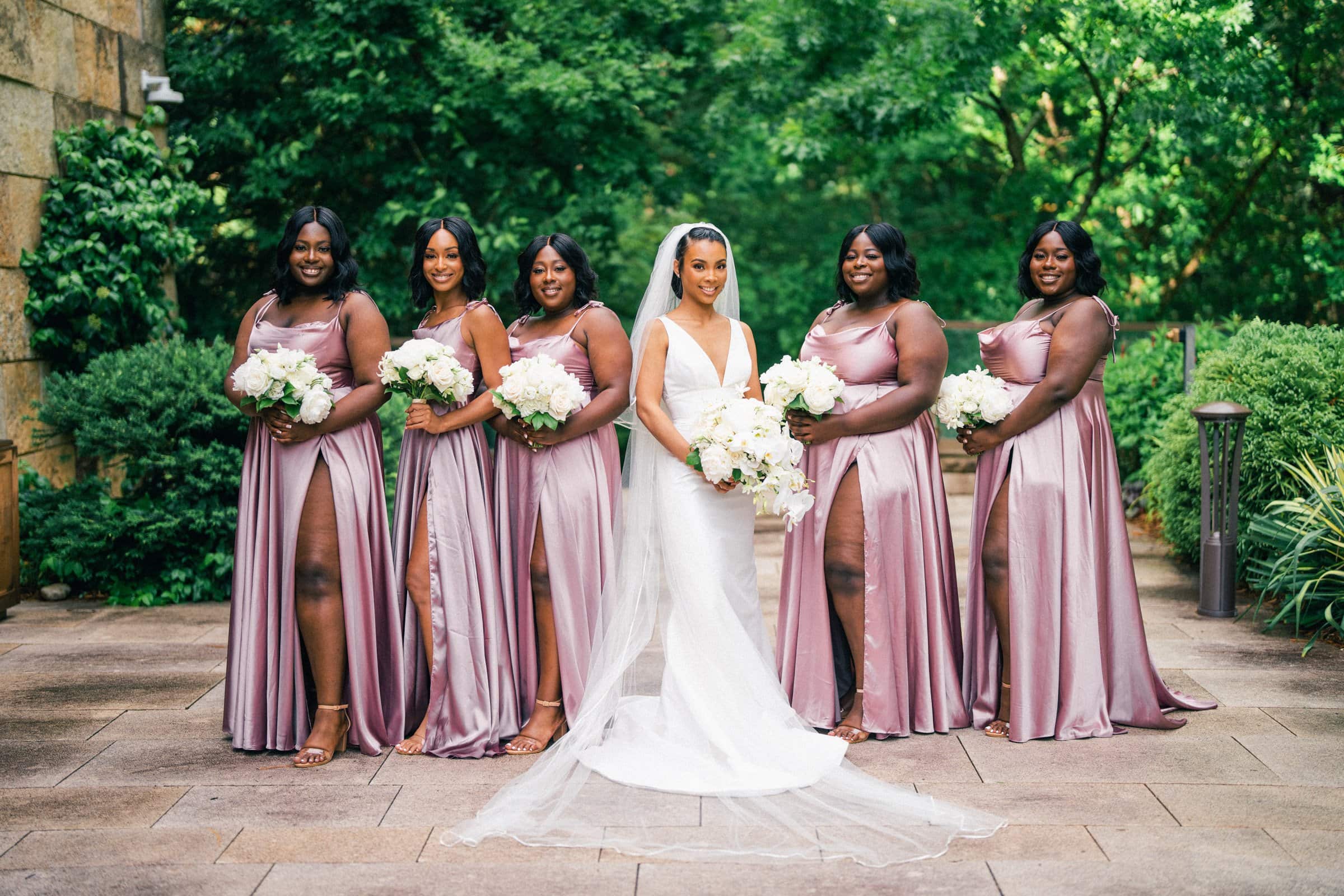 umstead wedding photography - kayla & deandre A bride in a white gown and veil stands with five bridesmaids in matching mauve dresses at this elegant Umstead wedding. They each hold bouquets of white flowers, posing outdoors amidst the lush greenery that frames this picturesque scene.