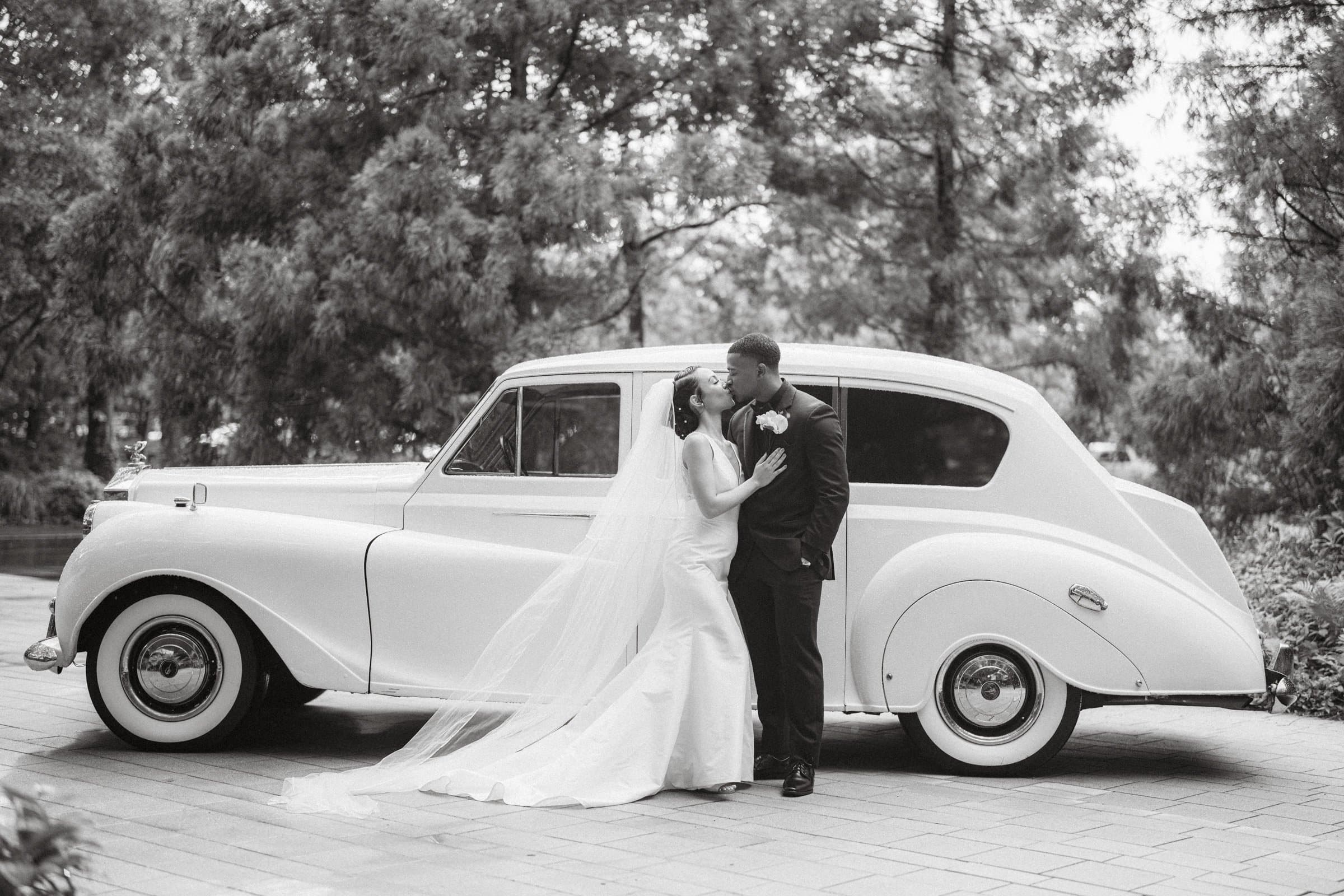 umstead wedding photography - kayla & deandre A bride and groom share a kiss in front of a classic white car at their Umstead wedding. The bride wears a long veil and an elegant dress, while the groom is in a dark suit. They stand on a paved path surrounded by trees, captured beautifully in black and white.