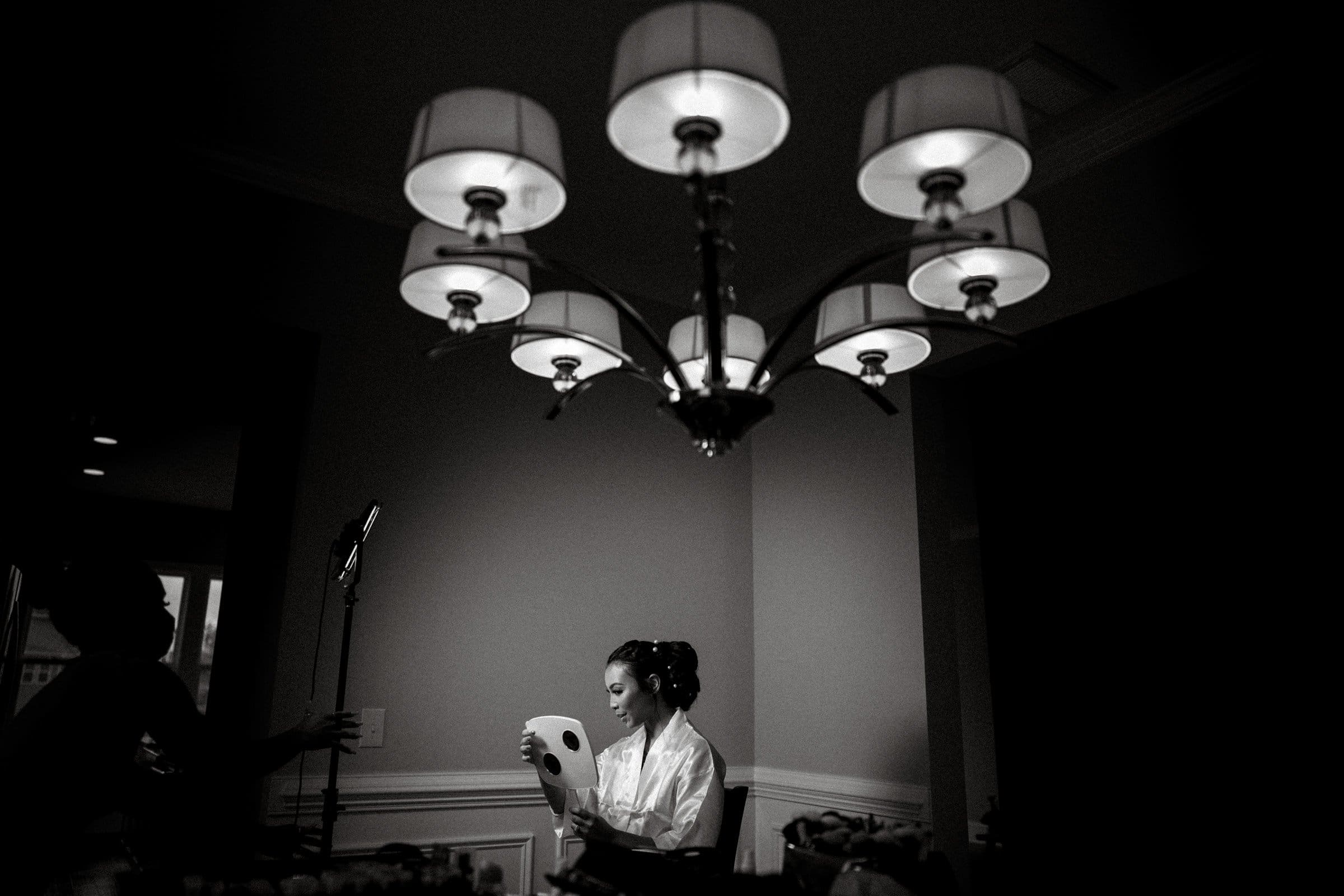 umstead wedding photography - kayla & deandre In the dimly lit room of the Umstead wedding, a person in a robe sits serenely under a large chandelier, gazing into a handheld mirror. Nearby, another individual stands focused, adjusting something on a tripod, adding to the calm and intimate atmosphere.
