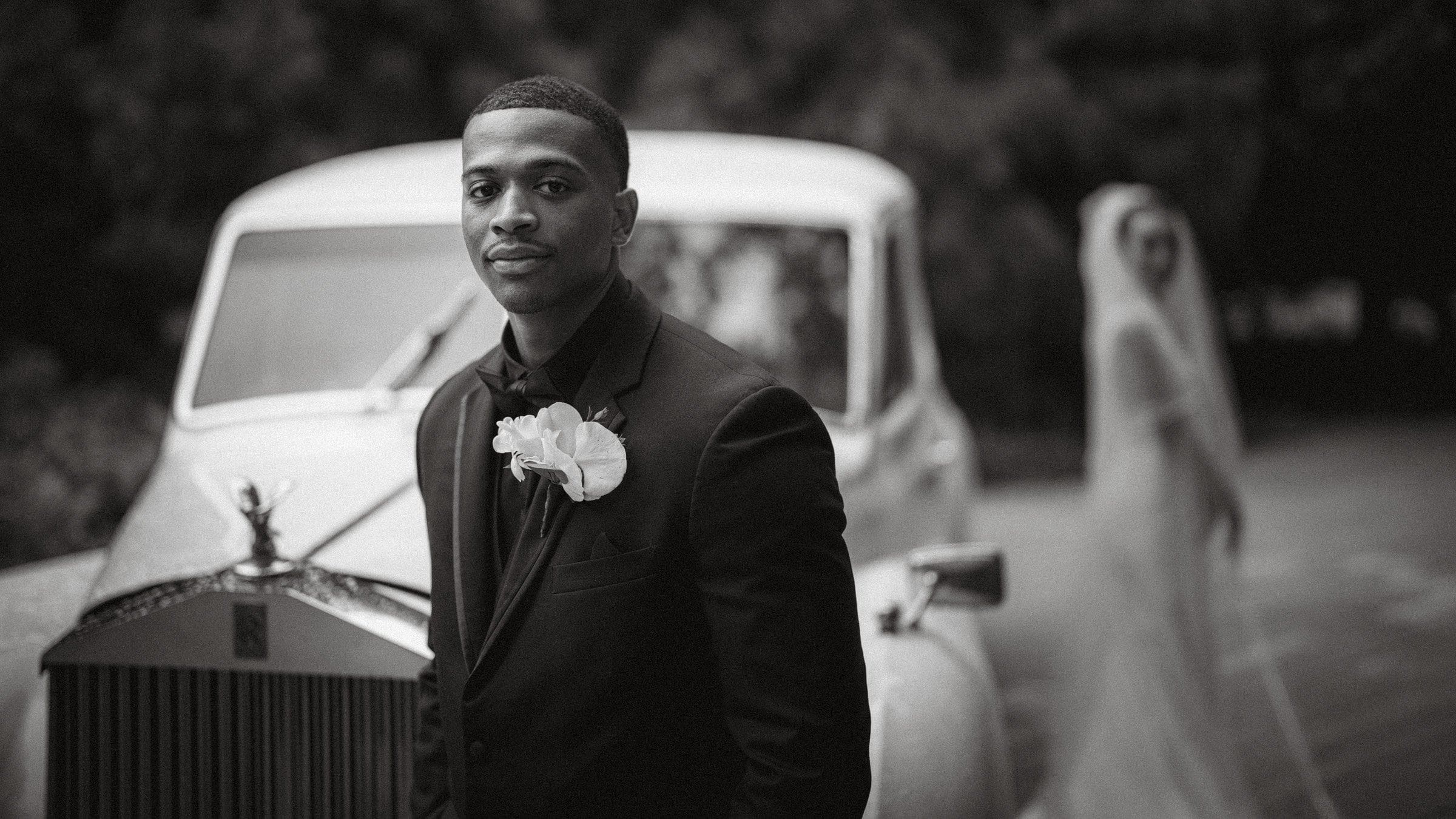 umstead wedding photography - kayla & deandre A man in a black suit with a white rose boutonniere stands proudly before a vintage car, capturing the timeless elegance of an Umstead wedding. In the softly out-of-focus background, a bride in her white gown and veil adds to the scene's classic allure. The entire image is beautifully rendered in black and white.