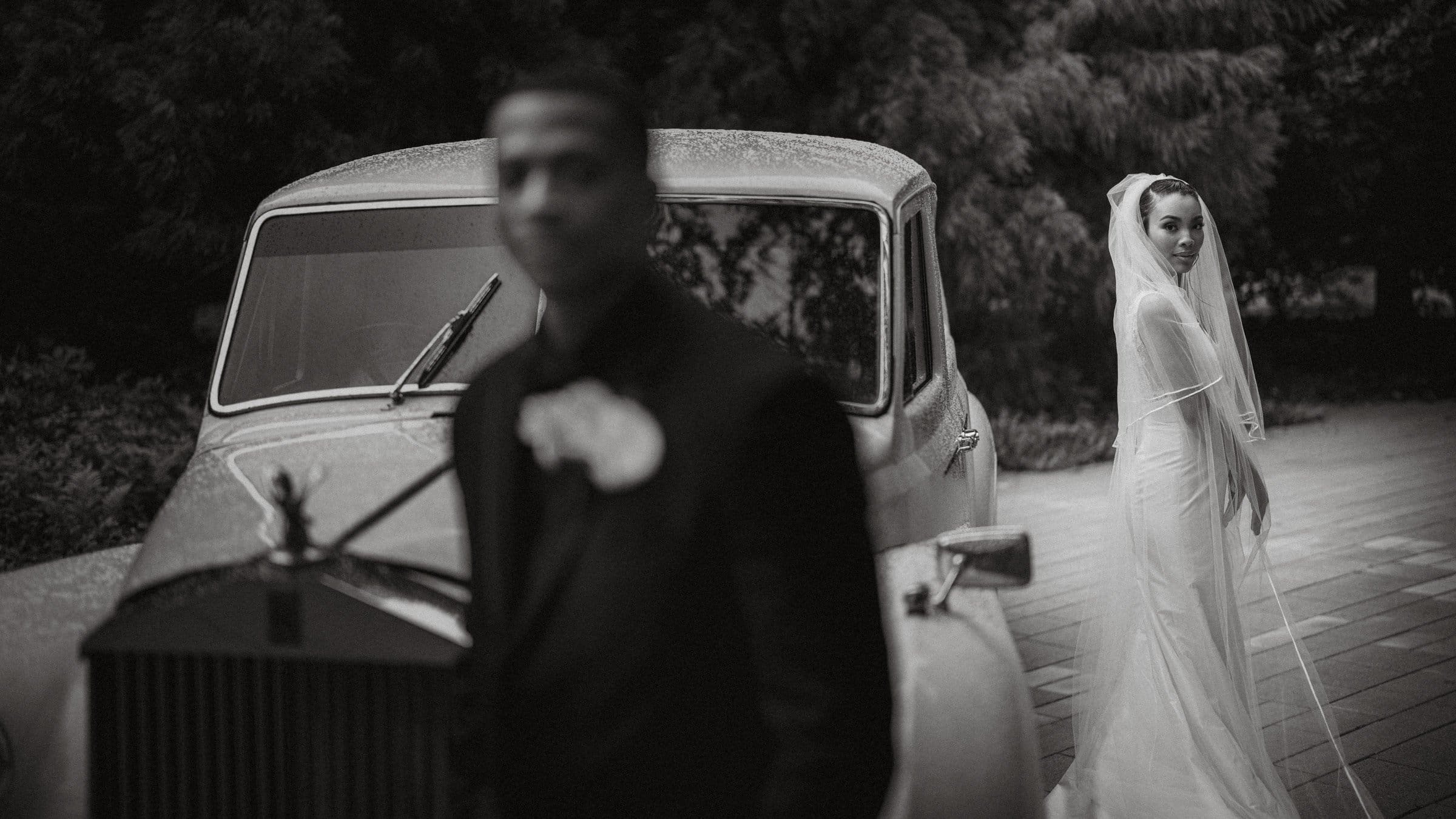 umstead wedding photography - kayla & deandre A vintage black and white photo captures an Umstead wedding scene with a bride in a long white gown and veil standing beside an old classic car. In the foreground, a man in a suit is blurred, adding depth to the image while lush greenery forms the backdrop.