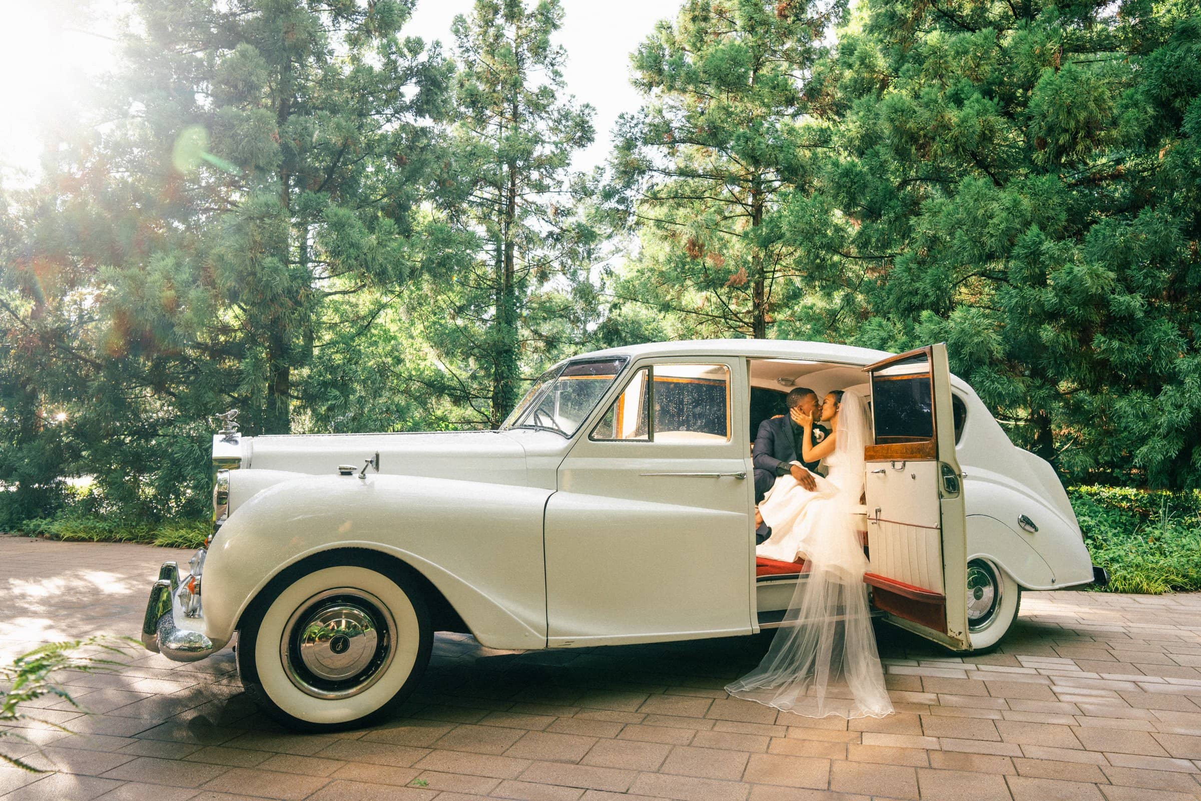 umstead wedding photography - kayla & deandre A bride and groom share a kiss inside a vintage white car with an open door at their enchanting Umstead wedding. The bride's veil flows elegantly outside the car, set on a sunlit pathway surrounded by tall, lush trees.