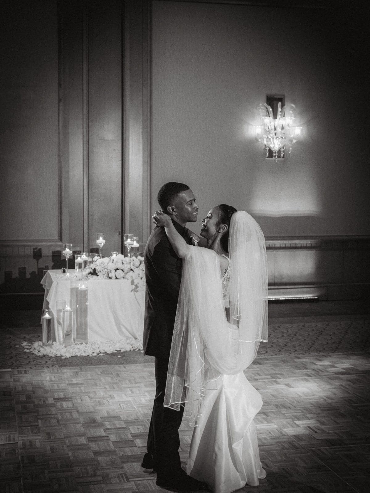 umstead wedding photography - kayla & deandre In a black and white photo of an Umstead wedding, a bride and groom share a dance in a dimly lit room. They embrace closely on the parquet floor, surrounded by elegant floral arrangements and the soft glow from a wall sconce.
