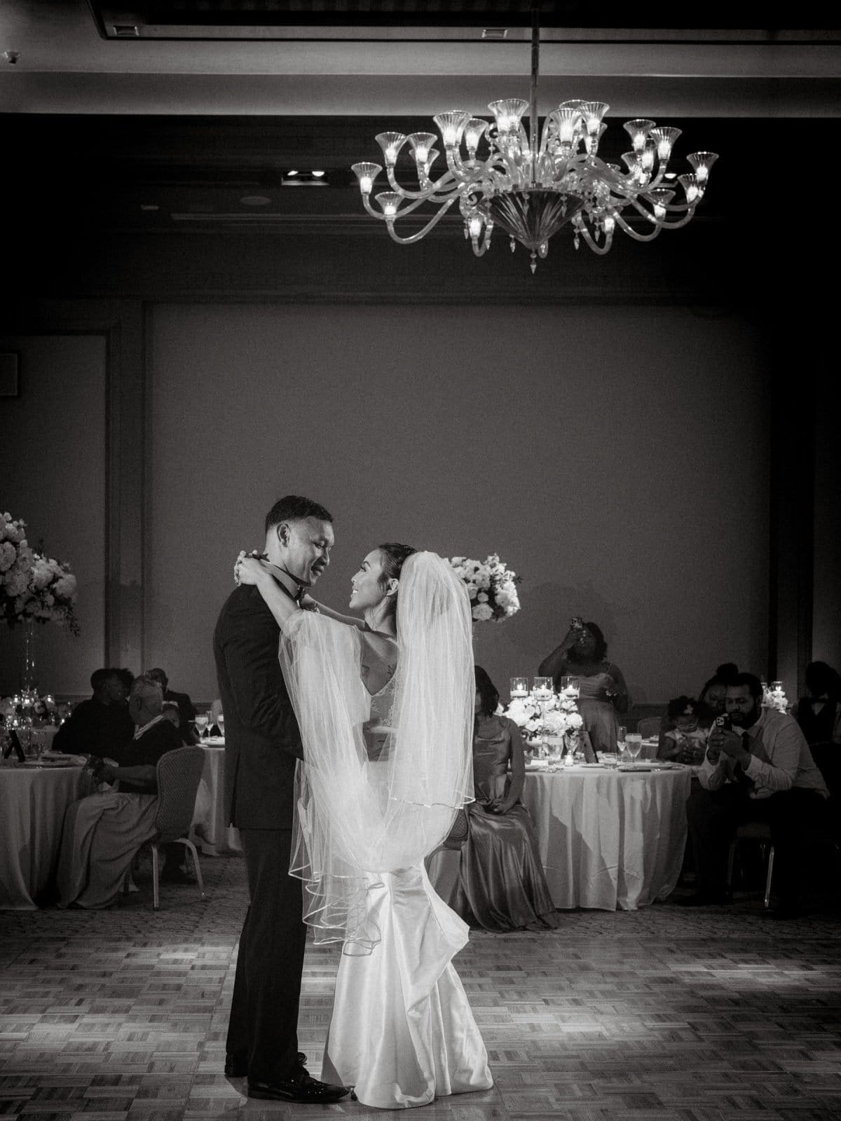 umstead wedding photography - kayla & deandre At their Umstead wedding, the bride and groom share their first dance under a chandelier in a dimly lit reception hall. Guests seated at decorated tables watch and take photos. The bride wears a veil and long dress, while the groom is in a suit.