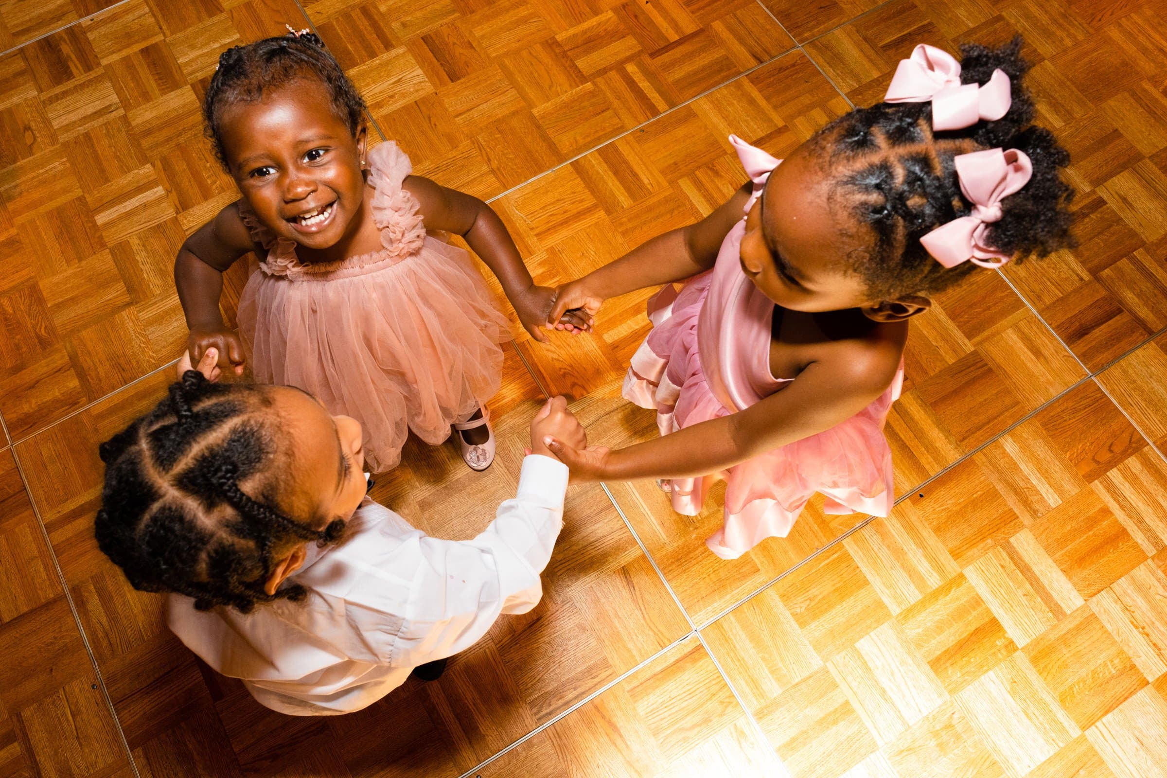 umstead wedding photography - kayla & deandre Three young children in formal attire hold hands in a circle on a wooden floor at an Umstead wedding. The two girls wear pink dresses with bows, and the boy is in a white shirt. They are smiling and appear to be enjoying a playful moment together.