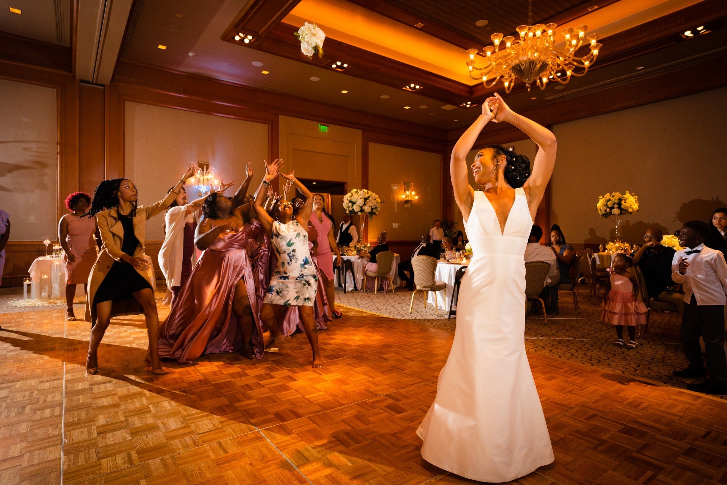 umstead wedding photography - kayla & deandre At an elegant Umstead wedding, a bride in a white gown prepares to toss her bouquet to eager guests in a warmly lit reception hall. The attendees, dressed in stylish attire, reach upwards with anticipation as chandeliers and floral arrangements add a festive touch.