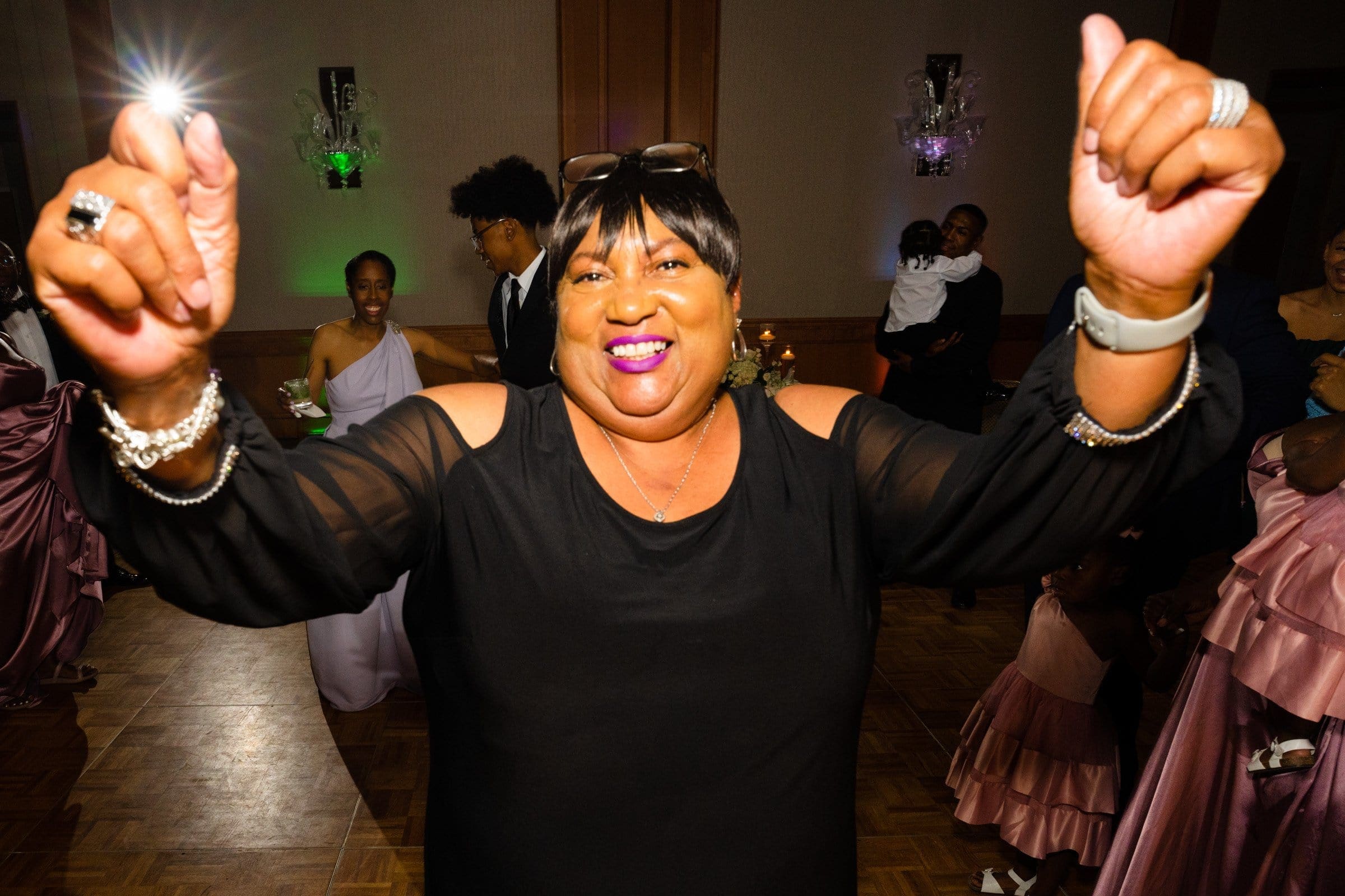 umstead wedding photography - kayla & deandre At an Umstead wedding, a joyful person with short hair, wearing a black dress, dances with raised hands at the festive event. People in elegant attire are seen in the background, including a person holding a baby. The scene is indoors with vibrant lighting.