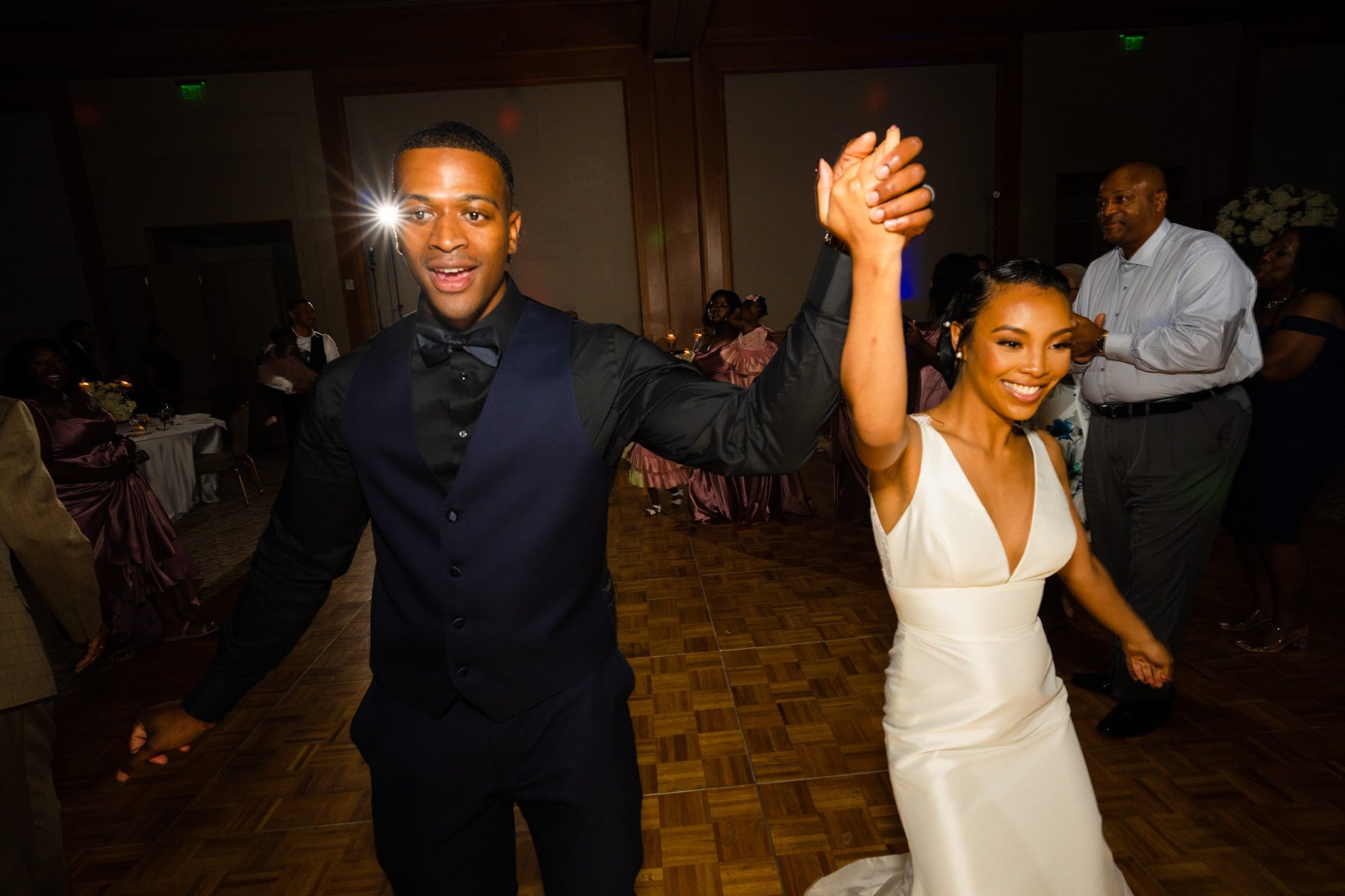 umstead wedding photography - kayla & deandre A joyful couple holds hands and dances at their Umstead wedding celebration. The woman wears a white gown, and the man is in a dark suit. Surrounded by guests on a wooden dance floor, a bright light shines in the background, adding to the festive atmosphere.