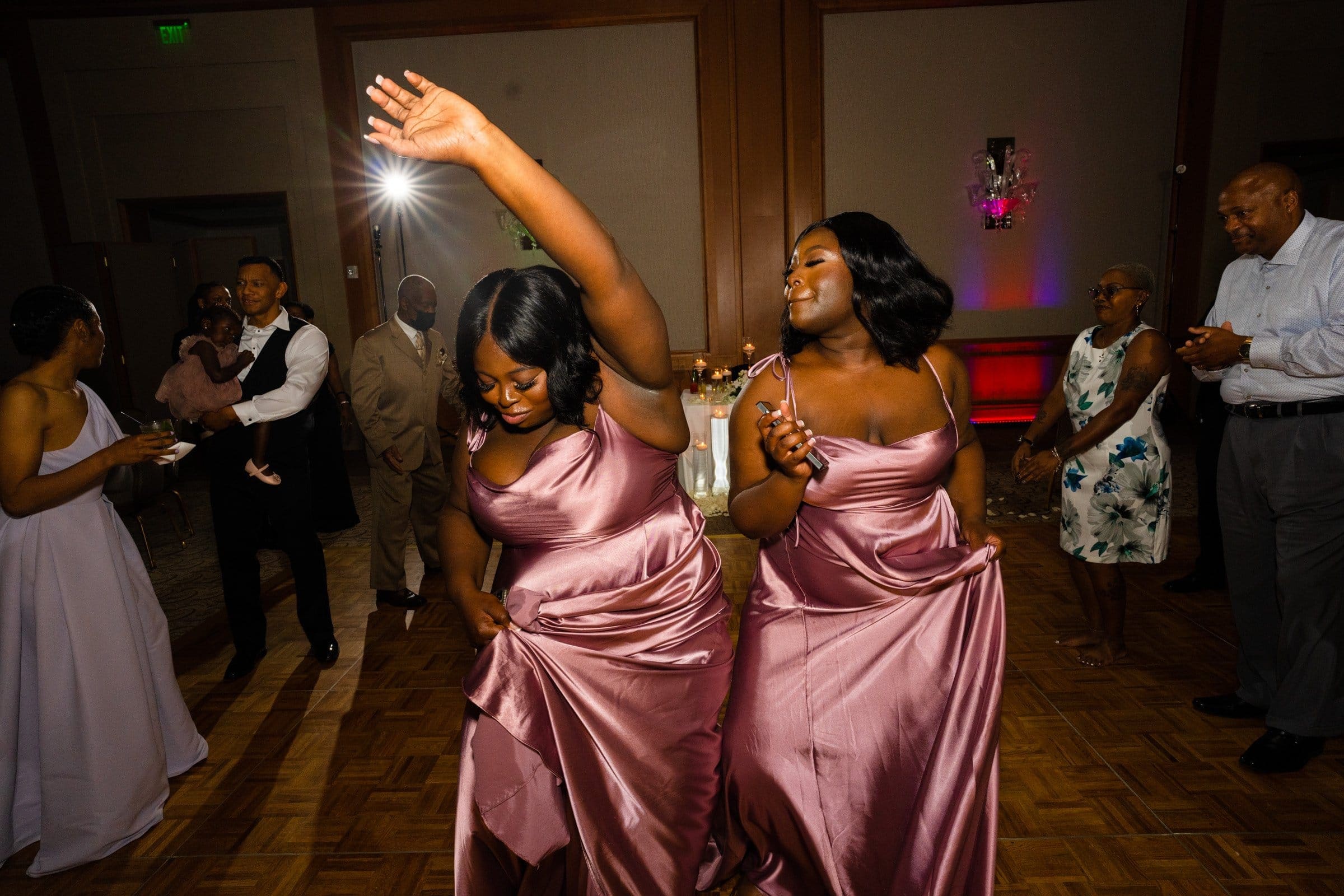 umstead wedding photography - kayla & deandre At the lively Umstead wedding, two women in matching mauve satin dresses joyfully dance on a wooden floor. Guests in the background watch and smile, as colorful lights illuminate the enchanting space.