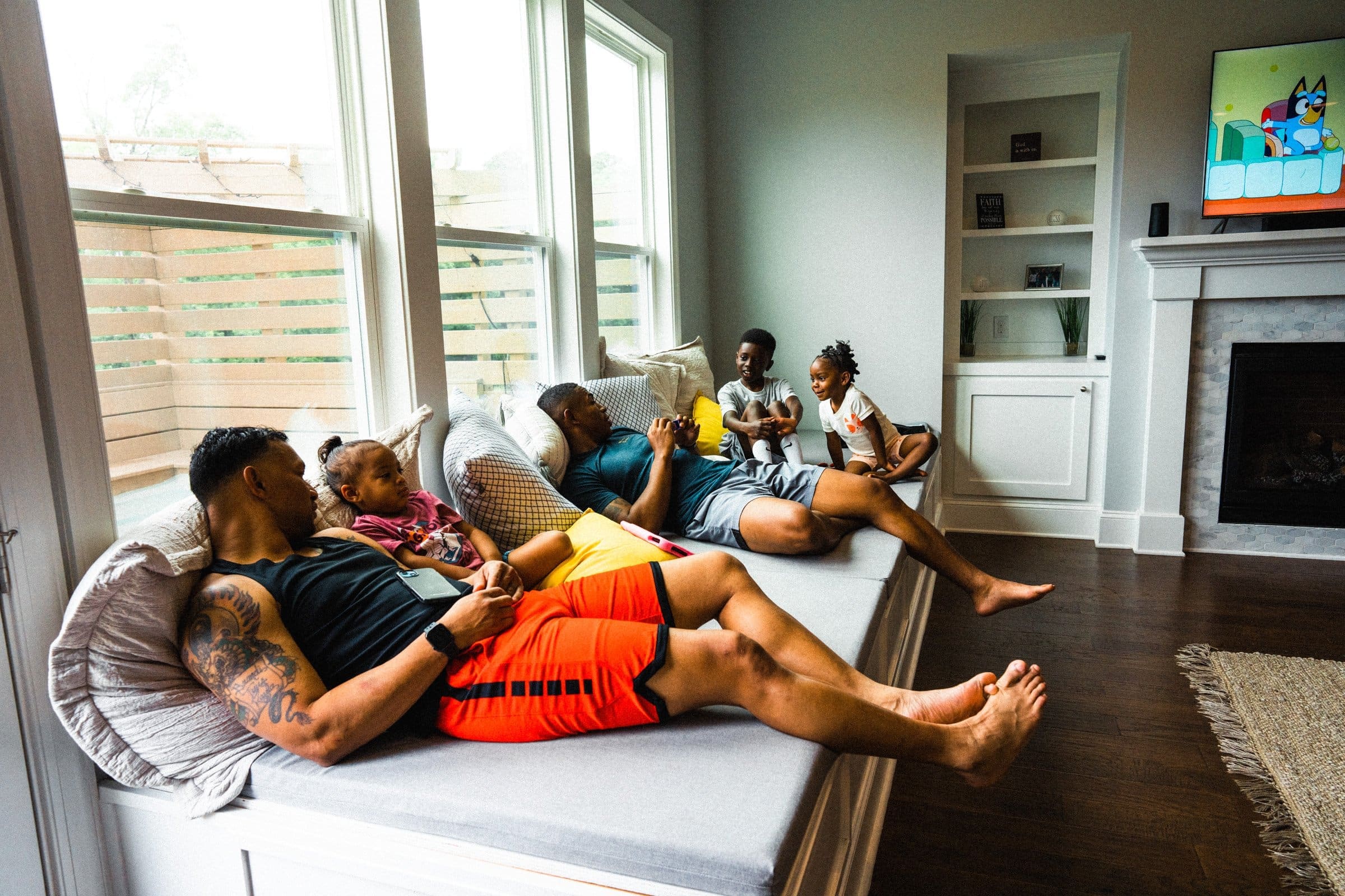 umstead wedding photography - kayla & deandre A group of people relax on a window seat in a living room, talking and reading. Two adults lounge in casual clothes while three children interact nearby, recounting memories of the recent Umstead wedding. A TV displays a cartoon, and natural light streams through the windows.