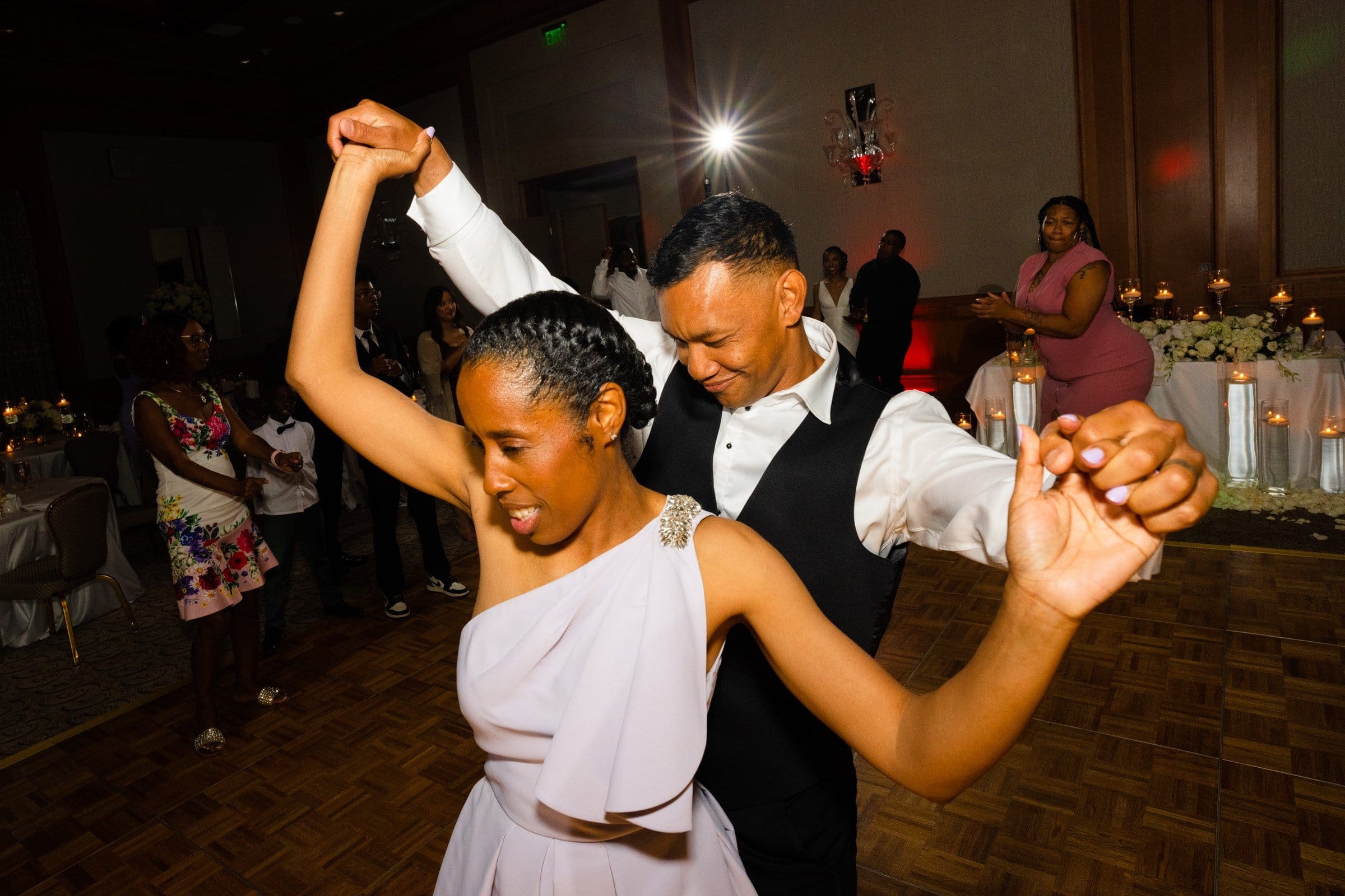 umstead wedding photography - kayla & deandre A couple dances joyfully at an Umstead wedding, with the woman in a white dress and the man in a white shirt and vest. Guests in elegant attire gather around, while tables set against the backdrop complete the enchanting indoor event.