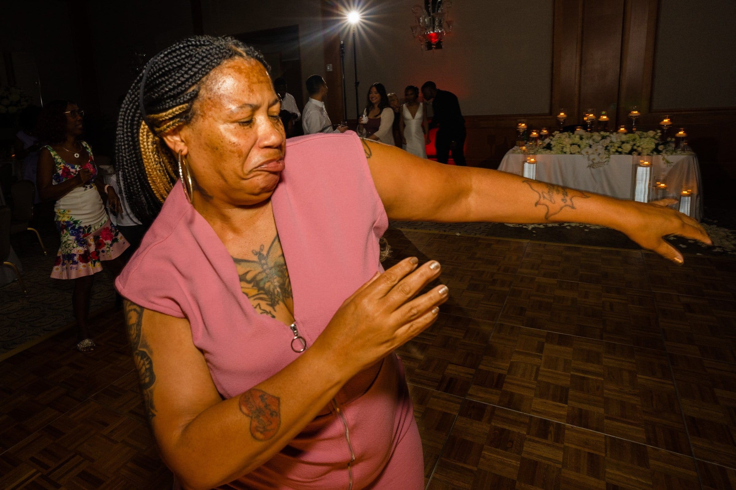 umstead wedding photography - kayla & deandre A person in a pink dress makes a dynamic dance move on the dimly lit dance floor of an Umstead wedding. In the background, guests gather near a table adorned with flowers and candles, suggesting a festive or celebratory event.