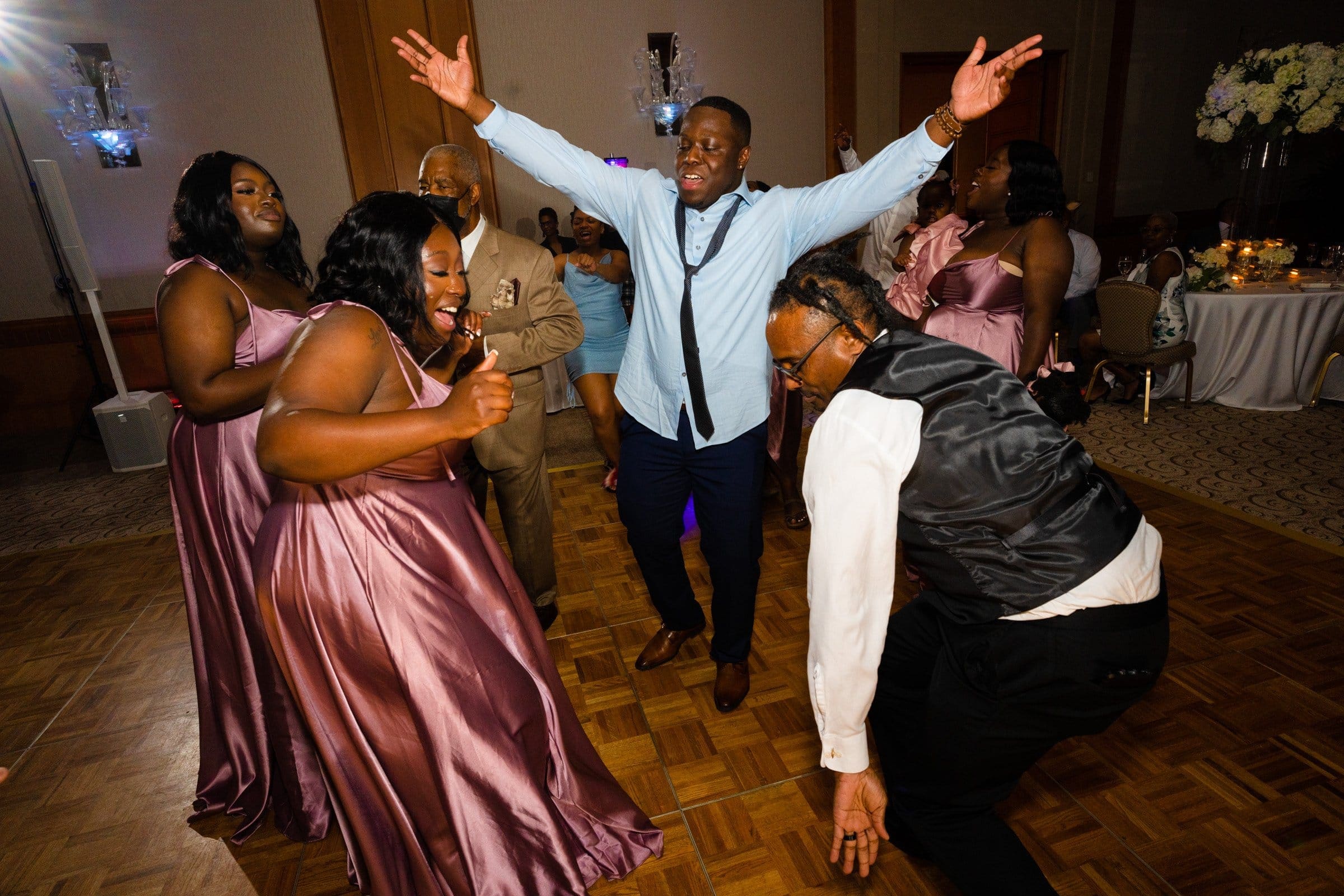 umstead wedding photography - kayla & deandre A joyful group of people dance together at an Umstead wedding celebration. They are dressed in formal attire, with the women in purple dresses and the men in suits. The room is warmly lit, and everyone's expressions show enjoyment and excitement.