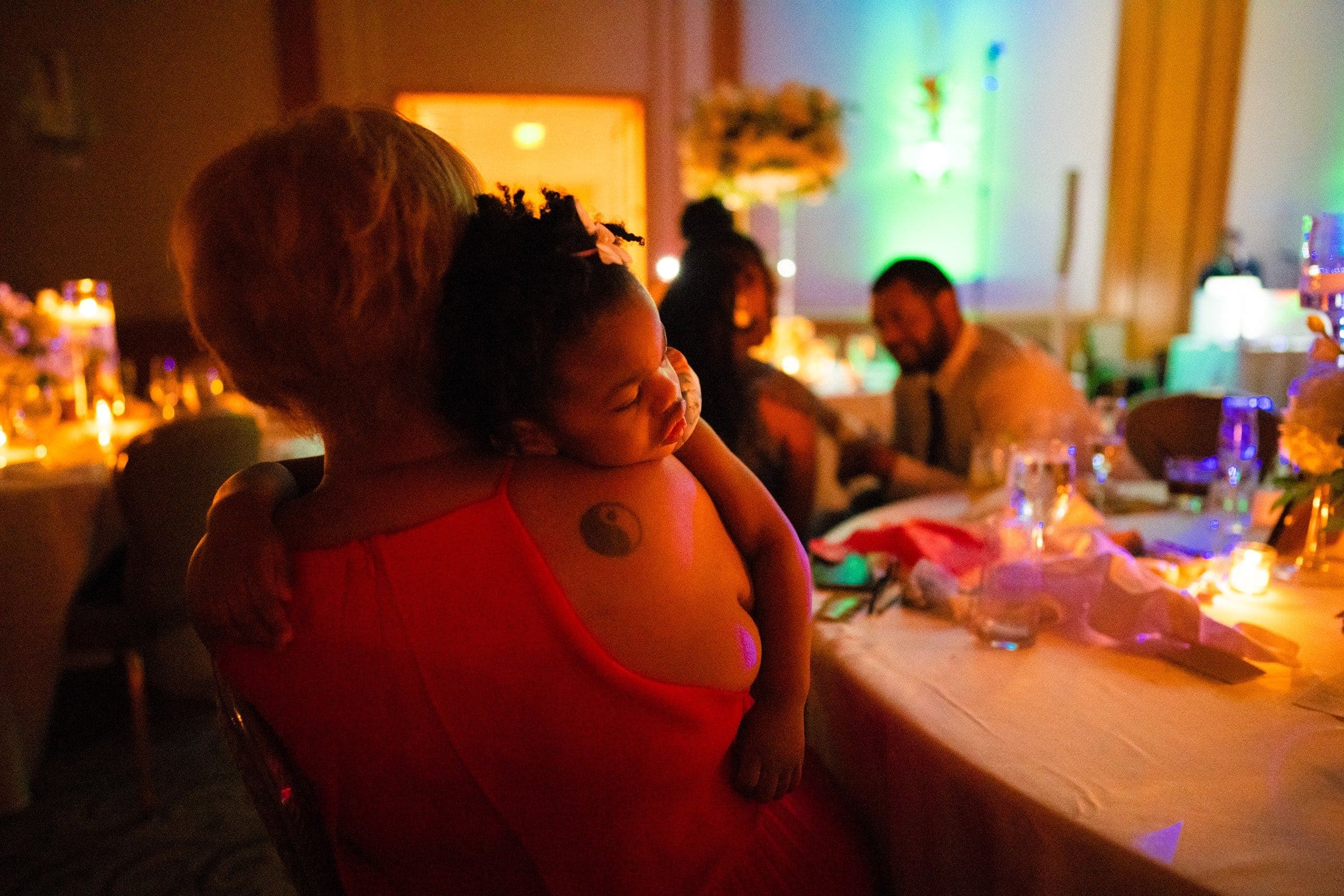 umstead wedding photography - kayla & deandre At an Umstead wedding, a woman in a red dress sits at a table, cradling a sleeping child on her shoulder. The dimly lit event is warmed by candles and soft lighting, while other guests enjoy the intimate ambiance around nearby tables.