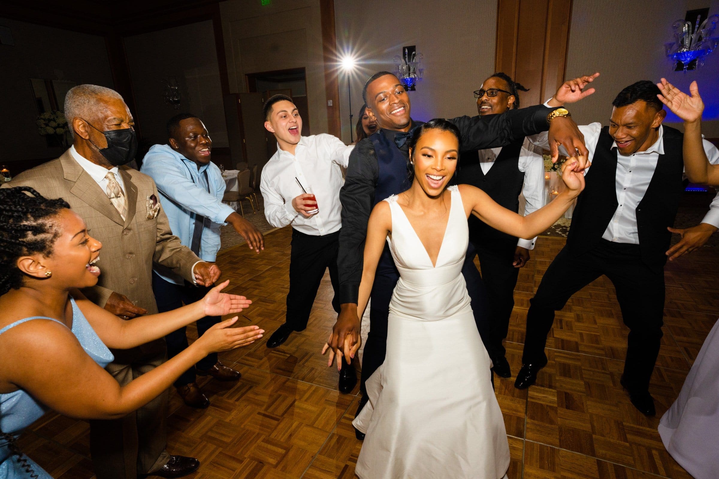 umstead wedding photography - kayla & deandre At their Umstead wedding, the bride in a white dress and the groom in a black tuxedo dance joyfully with guests. Women in blue dresses and men in suits clap and cheer on a wooden dance floor, illuminated by colorful lighting.