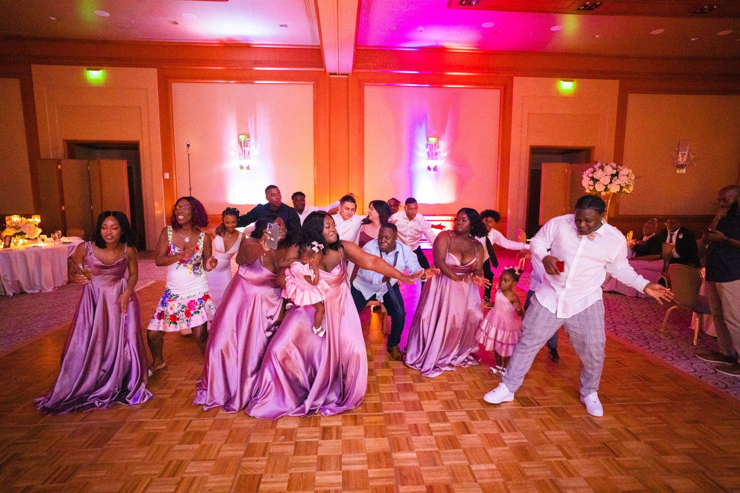 umstead wedding photography - kayla & deandre At an enchanting Umstead wedding, a group of people dressed in formal attire dance joyfully on a wooden floor. The room glows with purple and pink lighting. Women in matching lavender dresses and men in shirts and pants smile, reveling in the festive atmosphere.