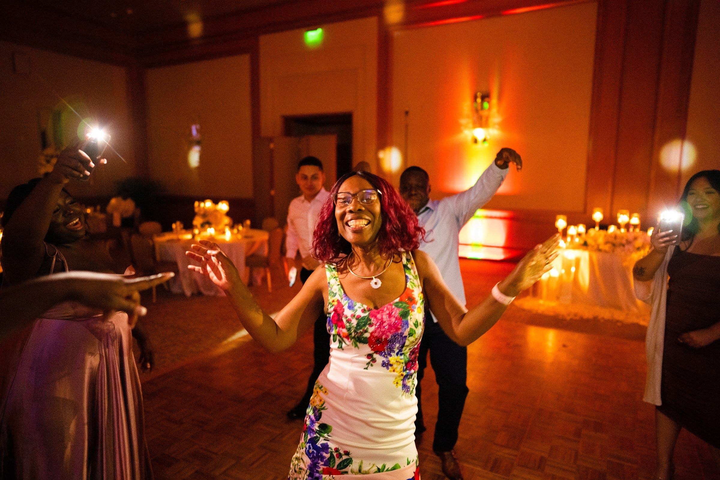 umstead wedding photography - kayla & deandre At an enchanting Umstead wedding, a woman in a floral dress happily dances on a wooden floor in the warmly lit room. Others around her are also dancing and snapping photos with their phones, while decorative candlelit tables add to the magic of the moment.