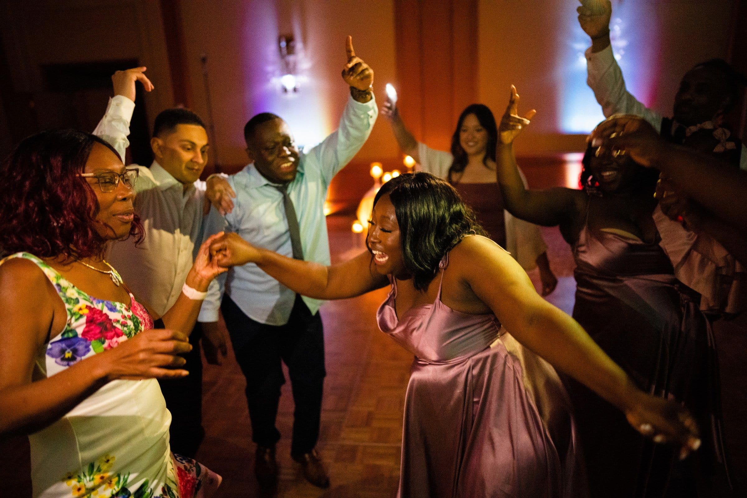 umstead wedding photography - kayla & deandre A joyful group of people dancing energetically at an indoor Umstead wedding. The scene is warmly lit, with individuals wearing elegant attire, including a woman in a floral dress and another in a purple dress, all reveling in the lively atmosphere.