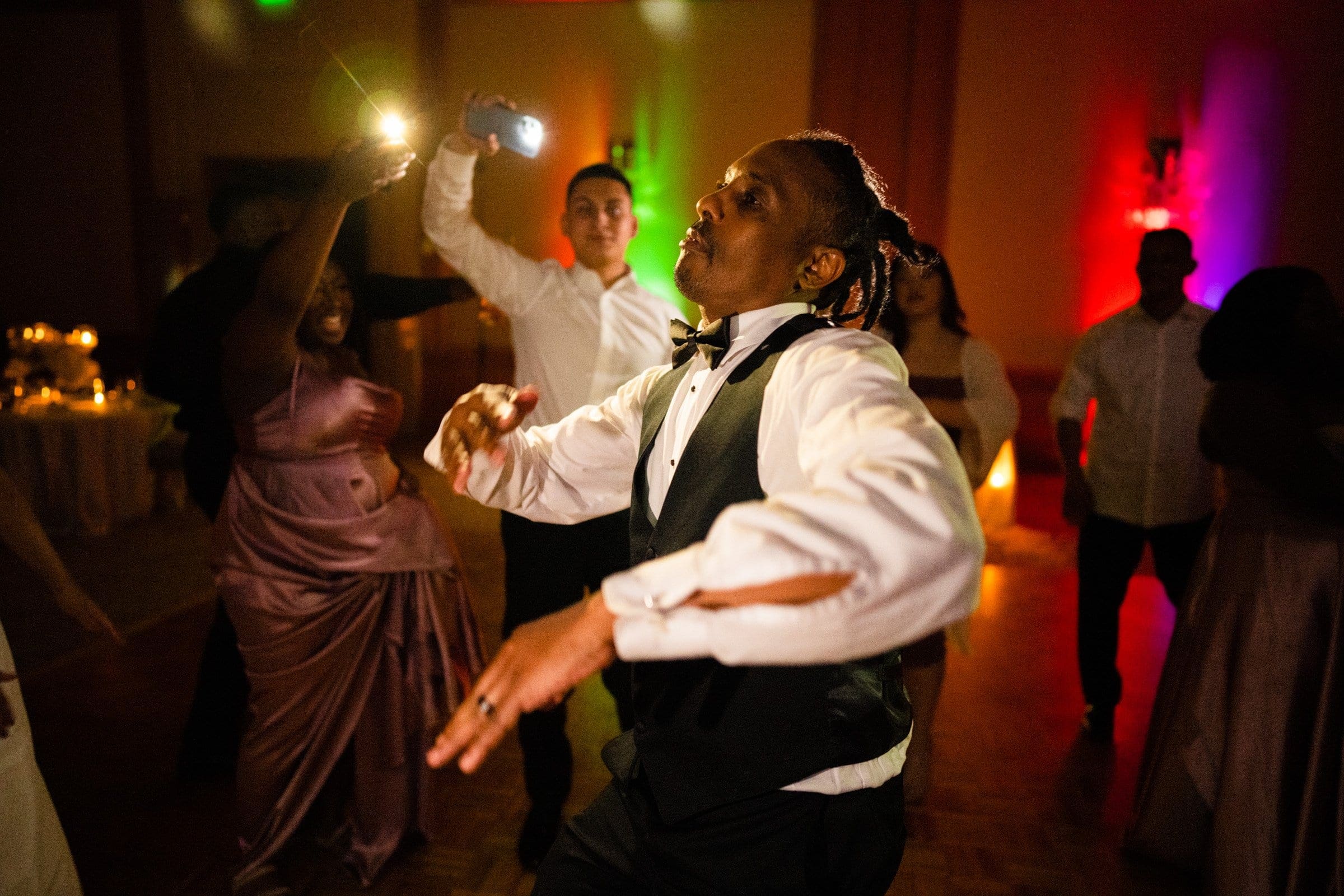 umstead wedding photography - kayla & deandre At an Umstead wedding, a man in a formal suit energetically dances at the party, surrounded by guests in elegant evening attire. Warm, colorful lights create a lively atmosphere on the dance floor.