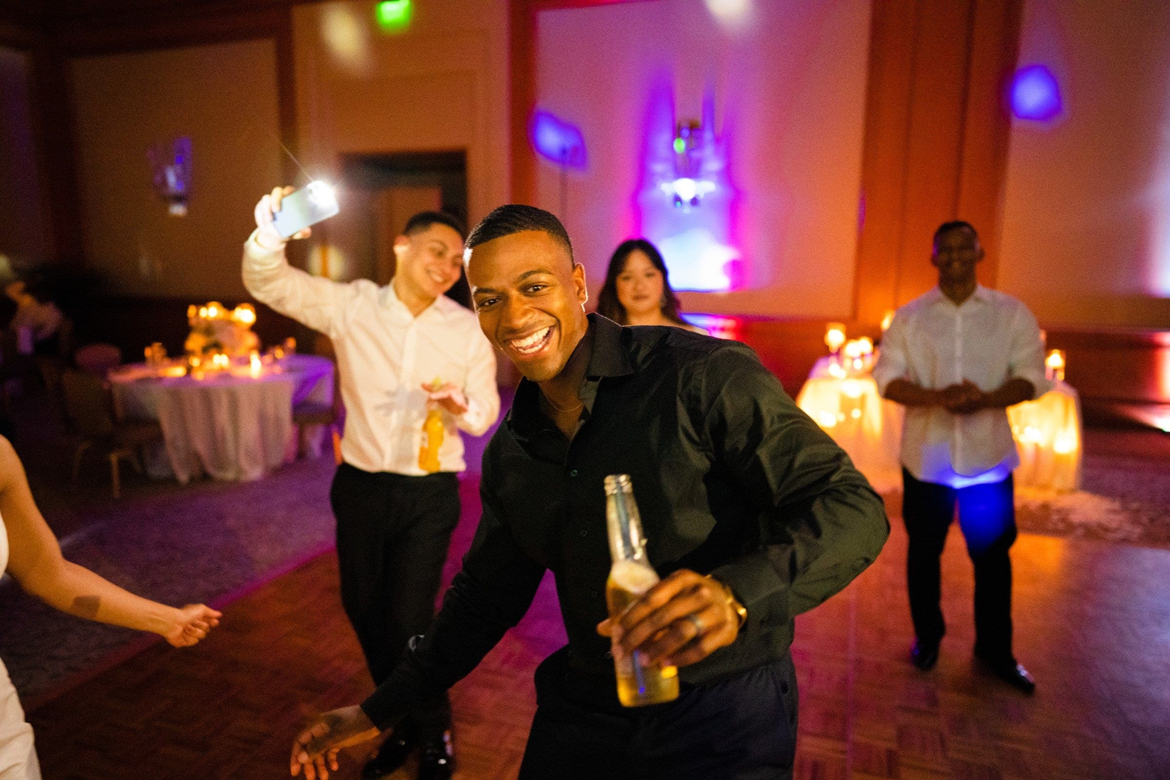 umstead wedding photography - kayla & deandre At an Umstead wedding, a lively group dances indoors. In the foreground, a man smiles, holding a bottle, while another captures the moment with a flash. The softly lit background is aglow with colorful lights and candle-decked tables, creating an enchanting atmosphere.