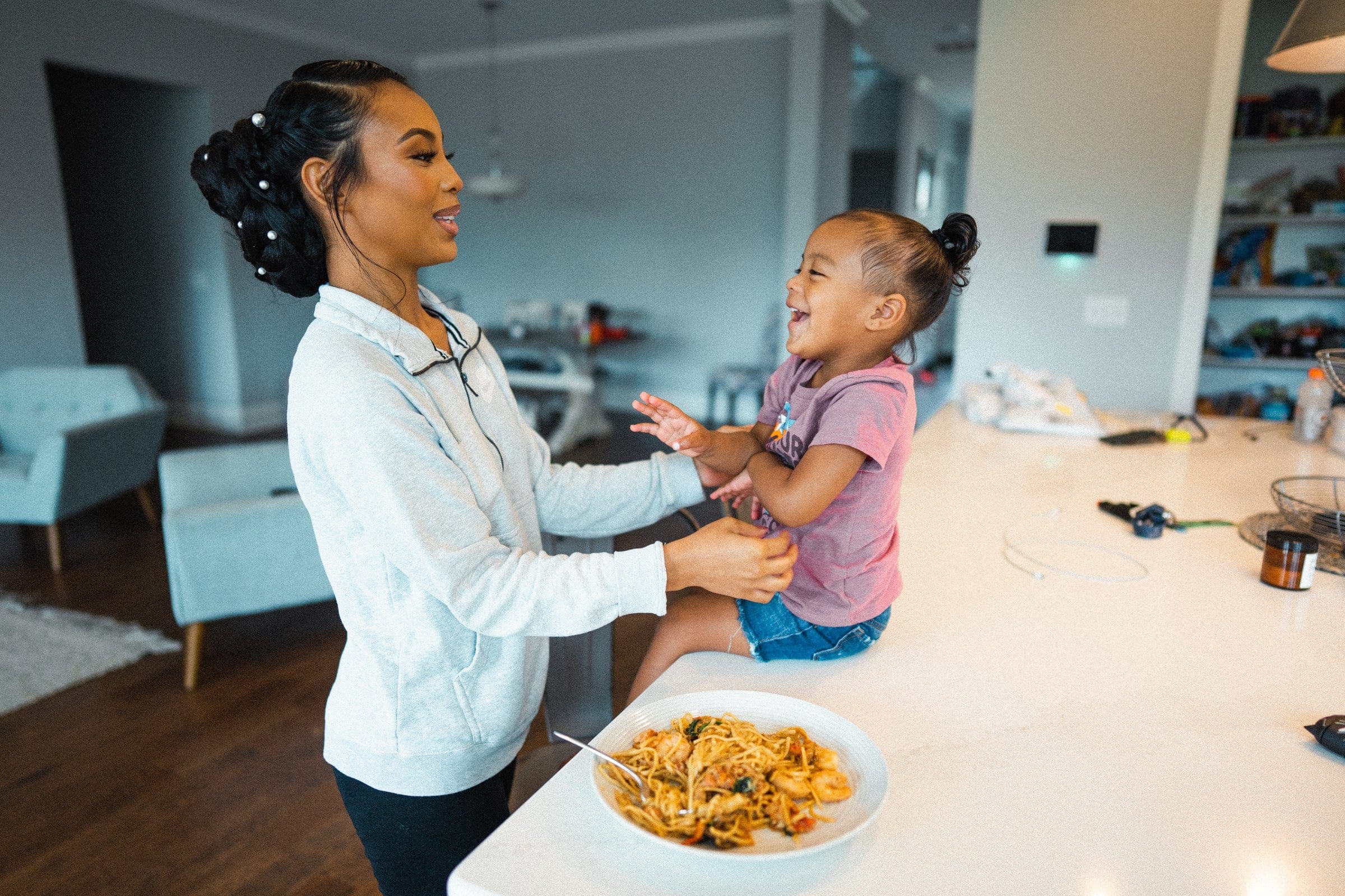 umstead wedding photography - kayla & deandre In a spacious, well-lit kitchen, where joy feels as tangible as the aroma of a family meal, a woman playfully interacts with a young girl perched on the counter. The girl beams in her pink shirt and denim shorts beside a plate of pasta, echoing the warmth of an intimate Umstead wedding.