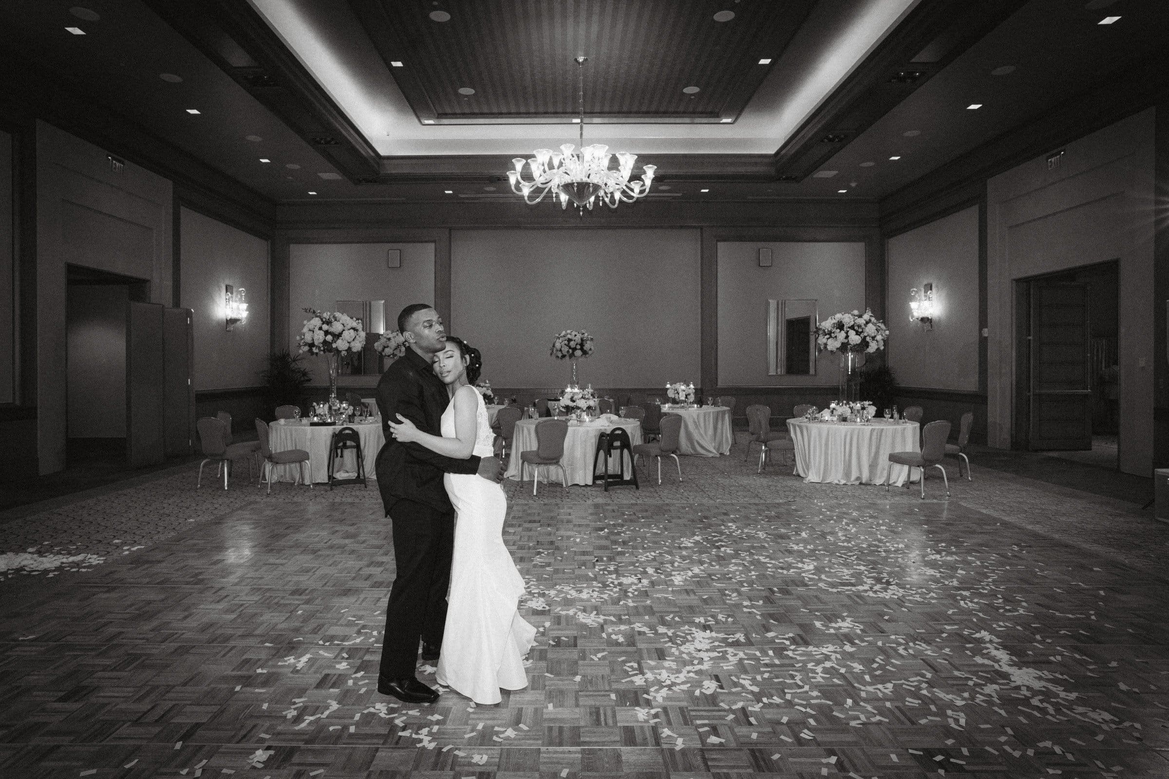 umstead wedding photography - kayla & deandre A couple dances alone on a confetti-strewn ballroom floor at their Umstead wedding. The room is elegantly decorated with floral arrangements and dim lighting, creating an enchanting atmosphere. A chandelier hangs from the ceiling, illuminating the couple in an intimate embrace.