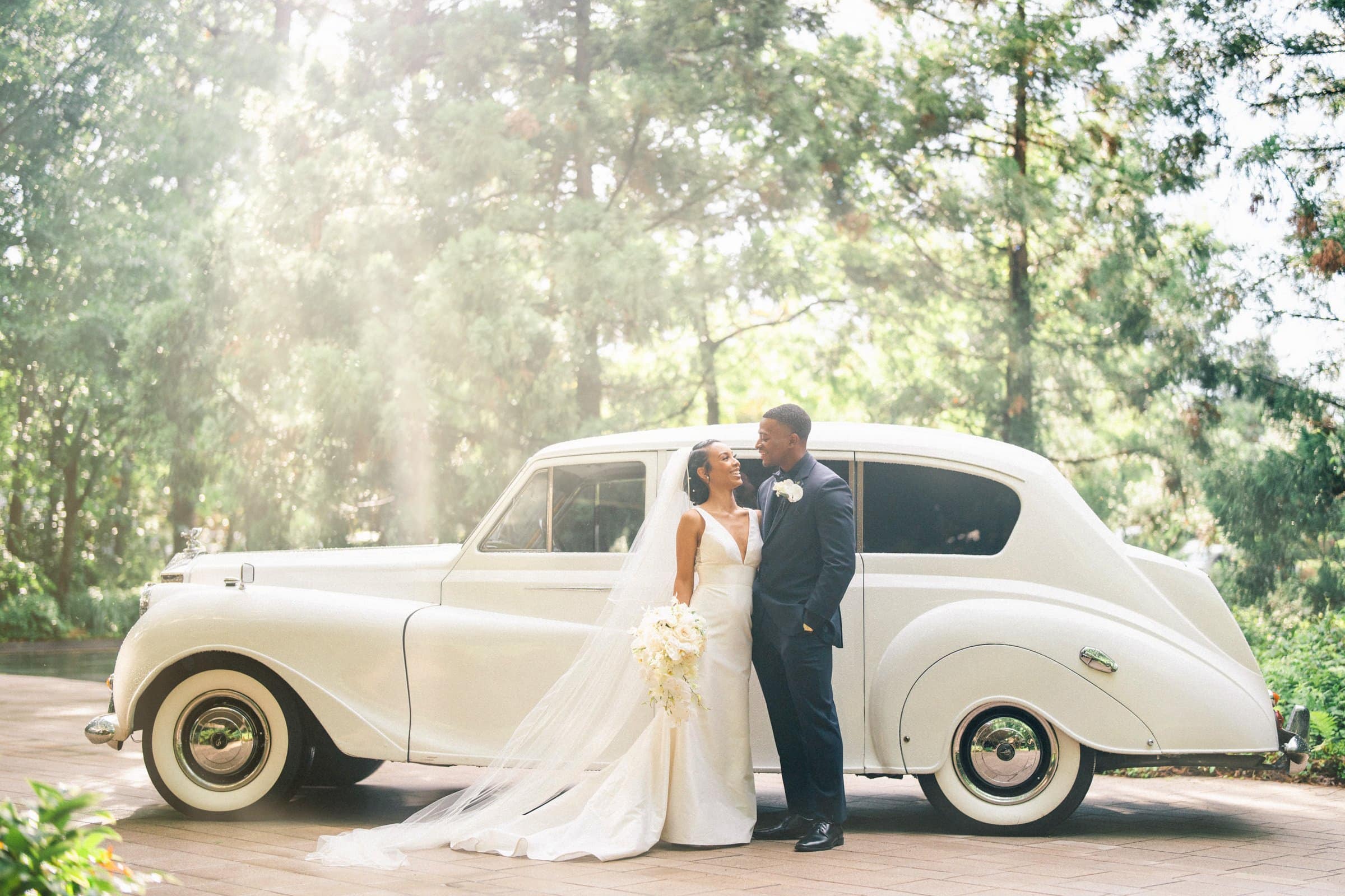 At their enchanting Umstead wedding, a bride and groom stand beside a classic white car in a lush, sunlit garden. The bride holds a bouquet and wears a long veil, while the groom is in a dark suit. They gaze lovingly at each other.
