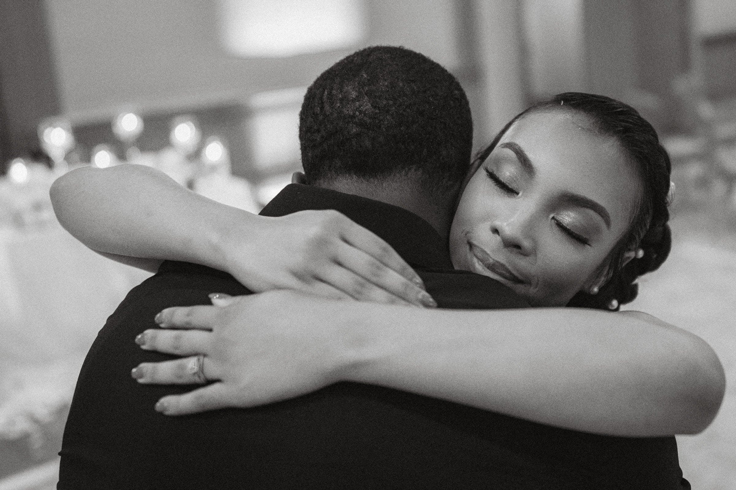 umstead wedding photography - kayla & deandre A black and white photo captures a tender moment at an Umstead wedding, with a woman embracing a man tightly. Her eyes are closed, and she appears content. The softly blurred background draws attention to their heartfelt embrace.