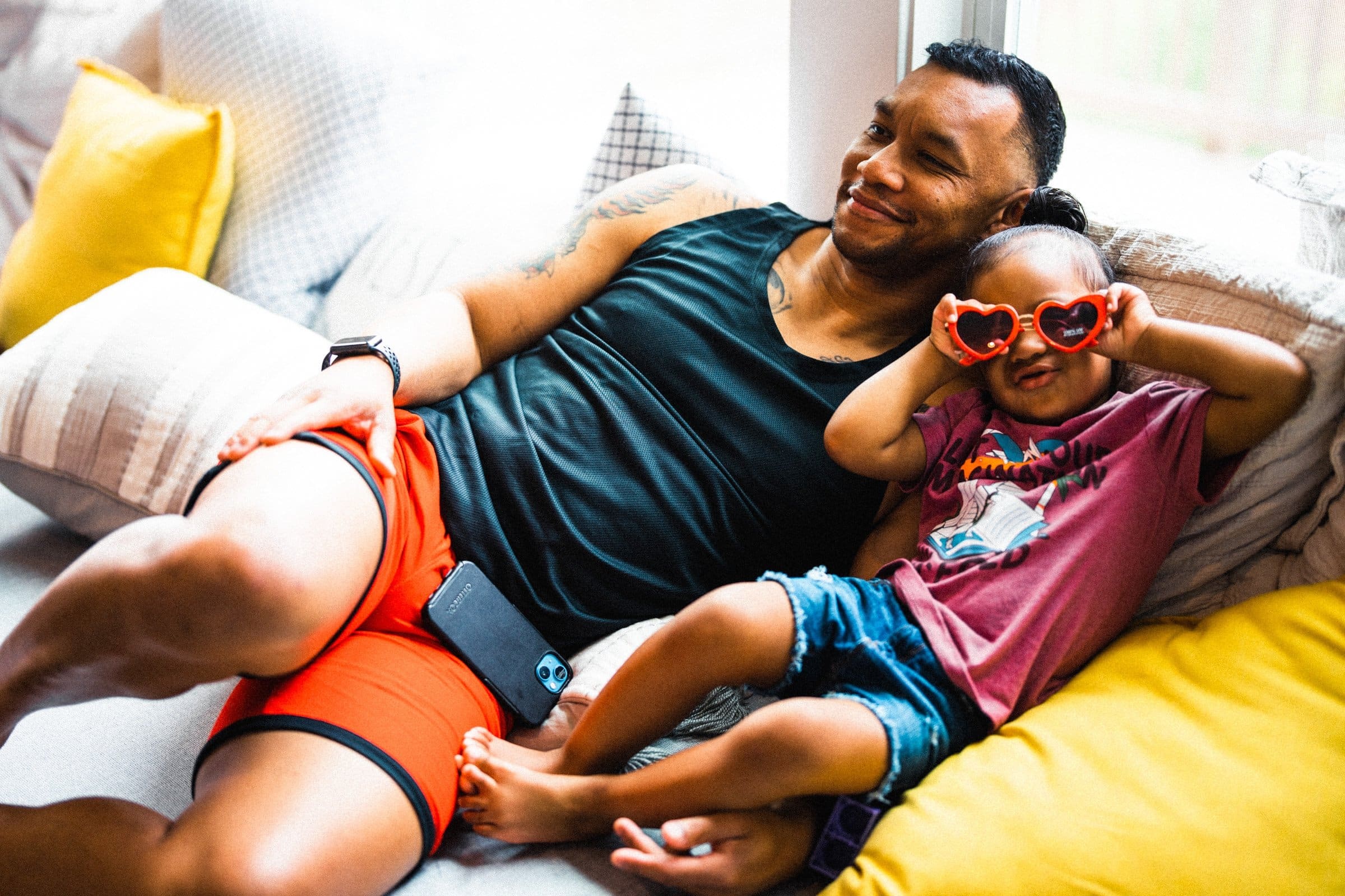 umstead wedding photography - kayla & deandre A man and a child relax on a sofa at the Umstead wedding. The man wears a black tank top and orange shorts, while the child sports a pink shirt with heart-shaped sunglasses. They both seem content, with the child playfully leaning on the man amidst the joyful celebration.