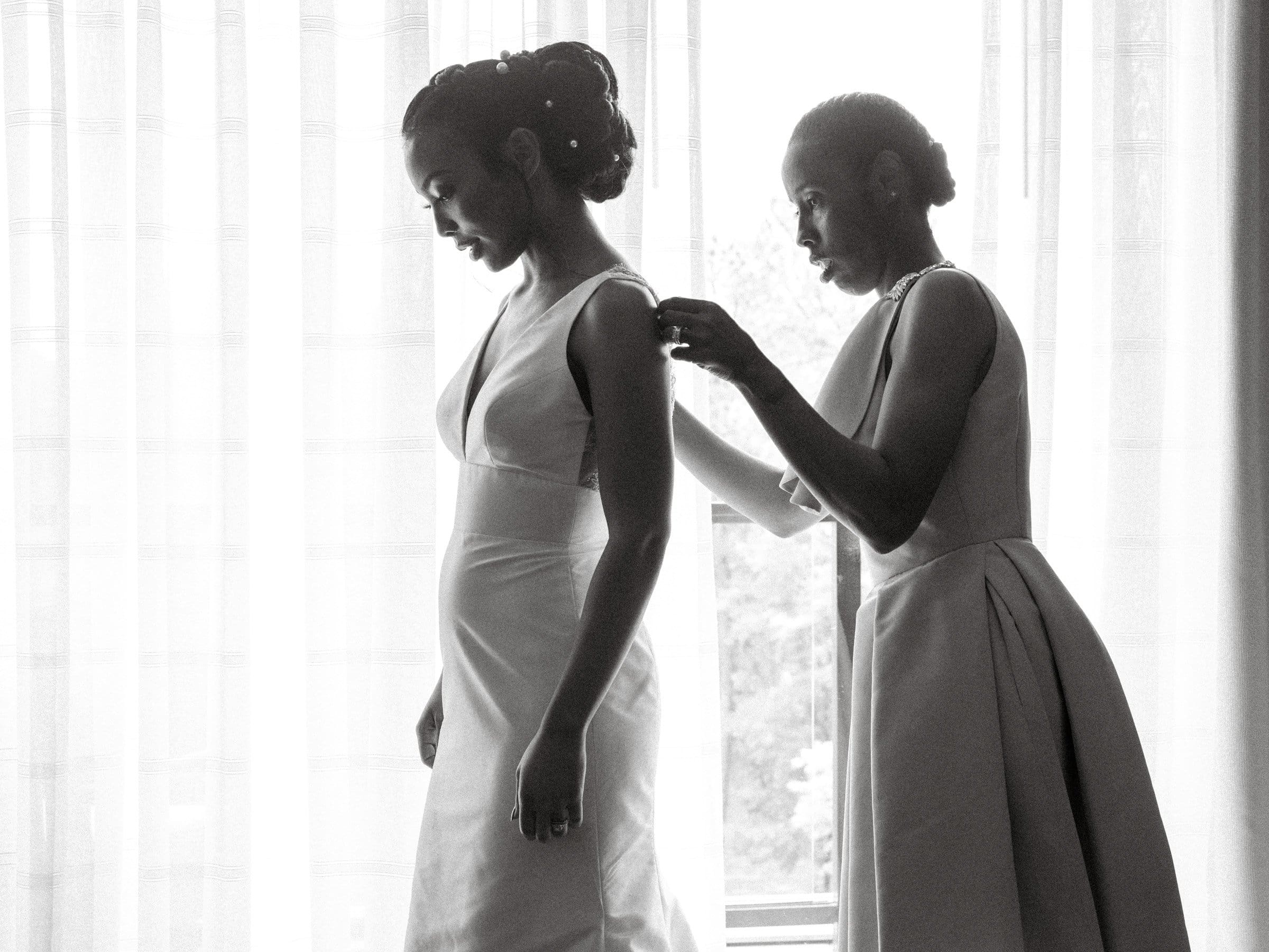 umstead wedding photography - kayla & deandre In a black-and-white photo of an elegant Umstead wedding, two women stand in a softly lit room. One wears a wedding dress while the other gently adjusts it near the window with sheer curtains, casting an intimate ambiance.