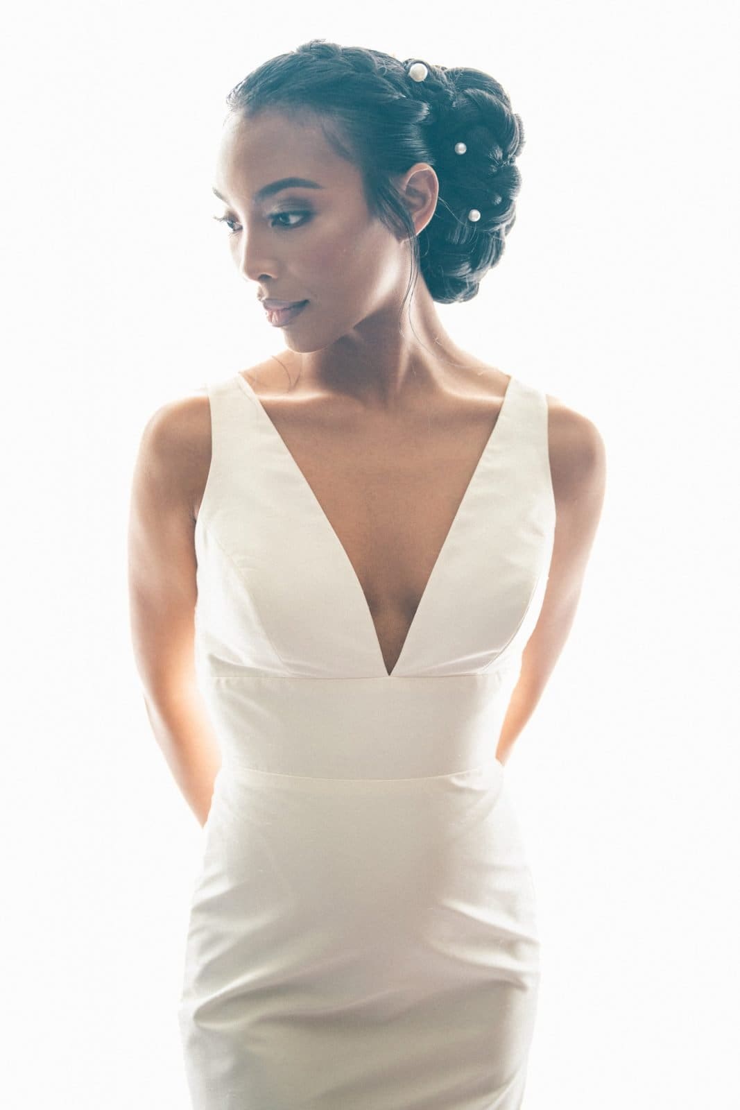 umstead wedding photography - kayla & deandre A woman in a white dress stands against a bright background, embodying an Umstead wedding's elegance. Her hair is styled up with decorative elements as she gazes to the side, hands behind her back. The dress features a deep neckline, with lighting that creates a soft, ethereal effect.