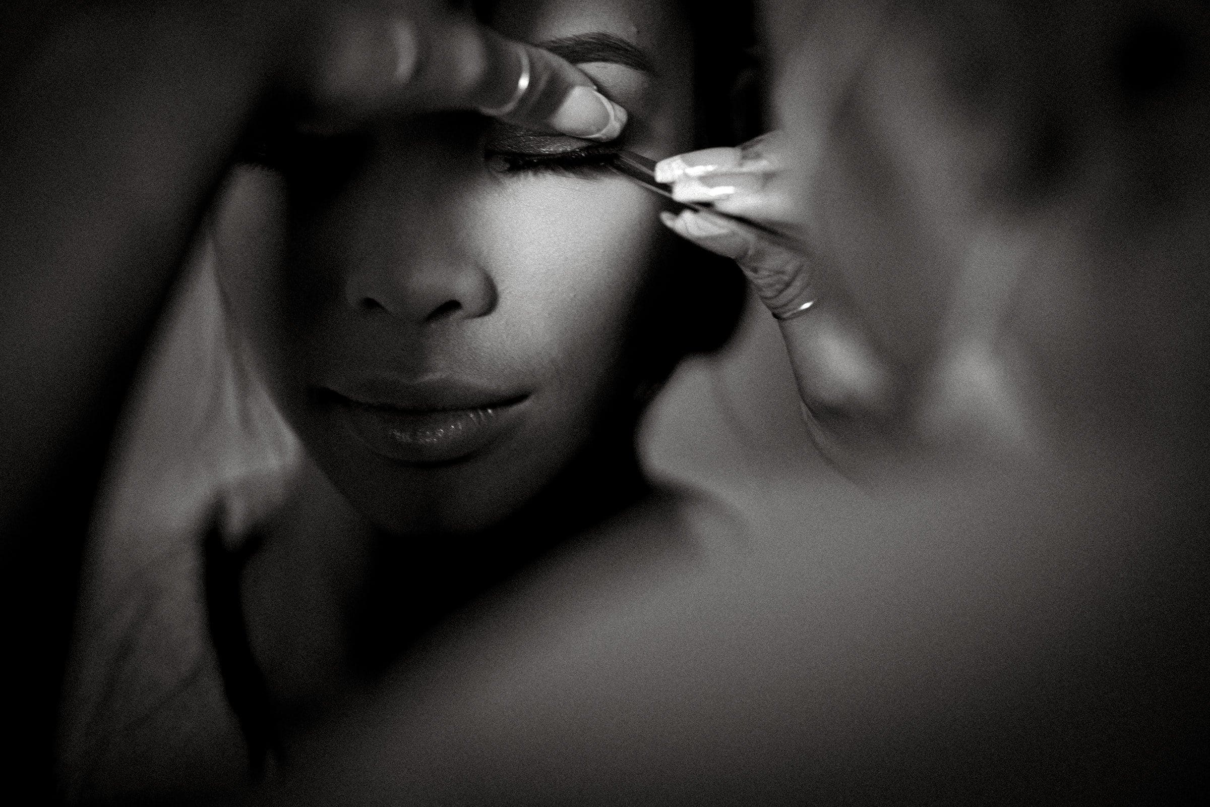 umstead wedding photography - kayla & deandre Close-up black and white image of a person having makeup applied to their eyes during an Umstead wedding. A hand gently holds an eye open while another applies eyeliner or mascara, creating a serene and focused atmosphere.