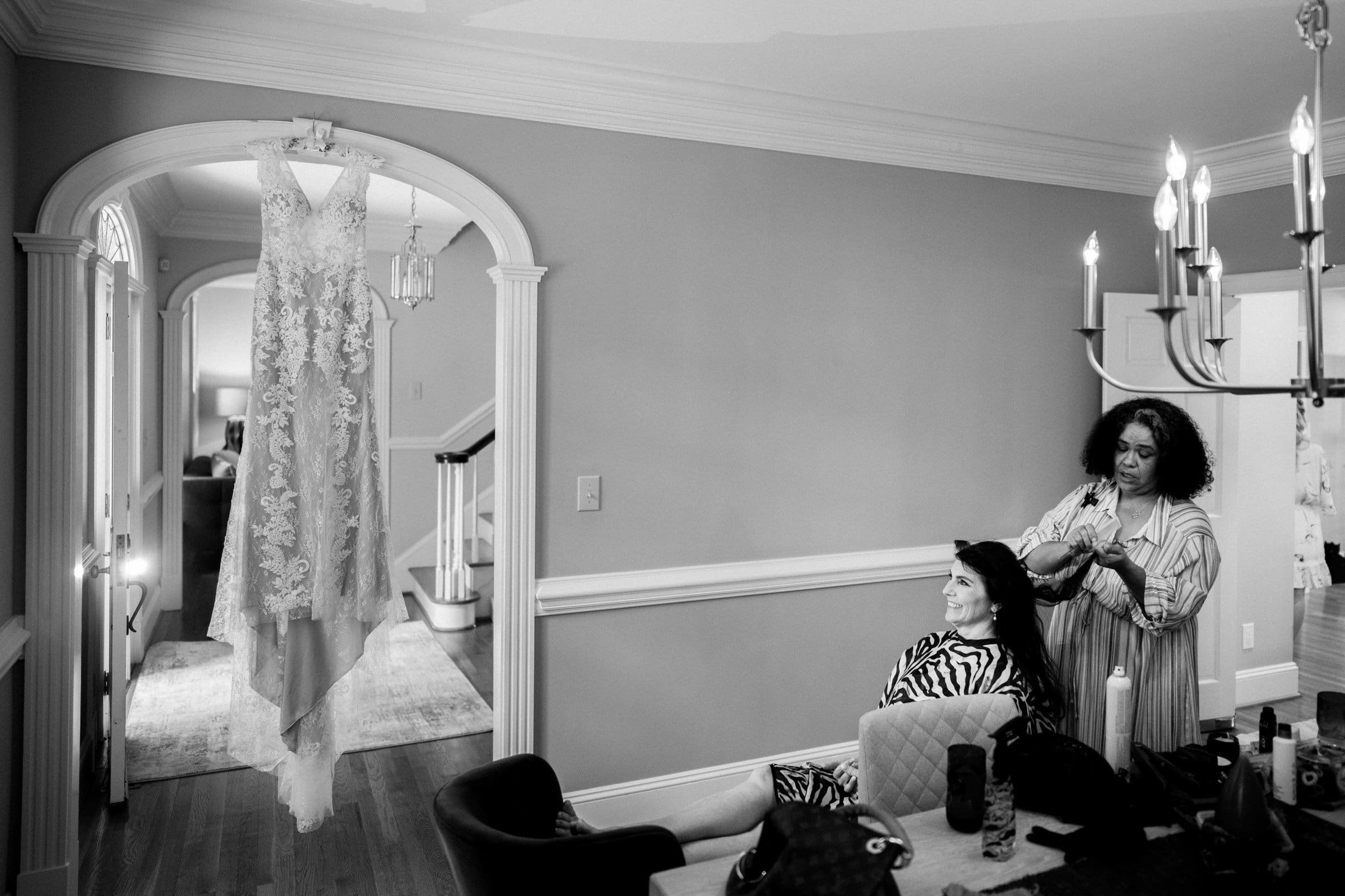 duke gardens wedding photography - ann & tim A black and white photo captures a bridal gown elegantly hanging in a doorway at Duke Gardens. A woman sits smiling as another styles her hair beneath a chandelier, the light-colored walls creating a warm and elegant atmosphere fitting for wedding photography.