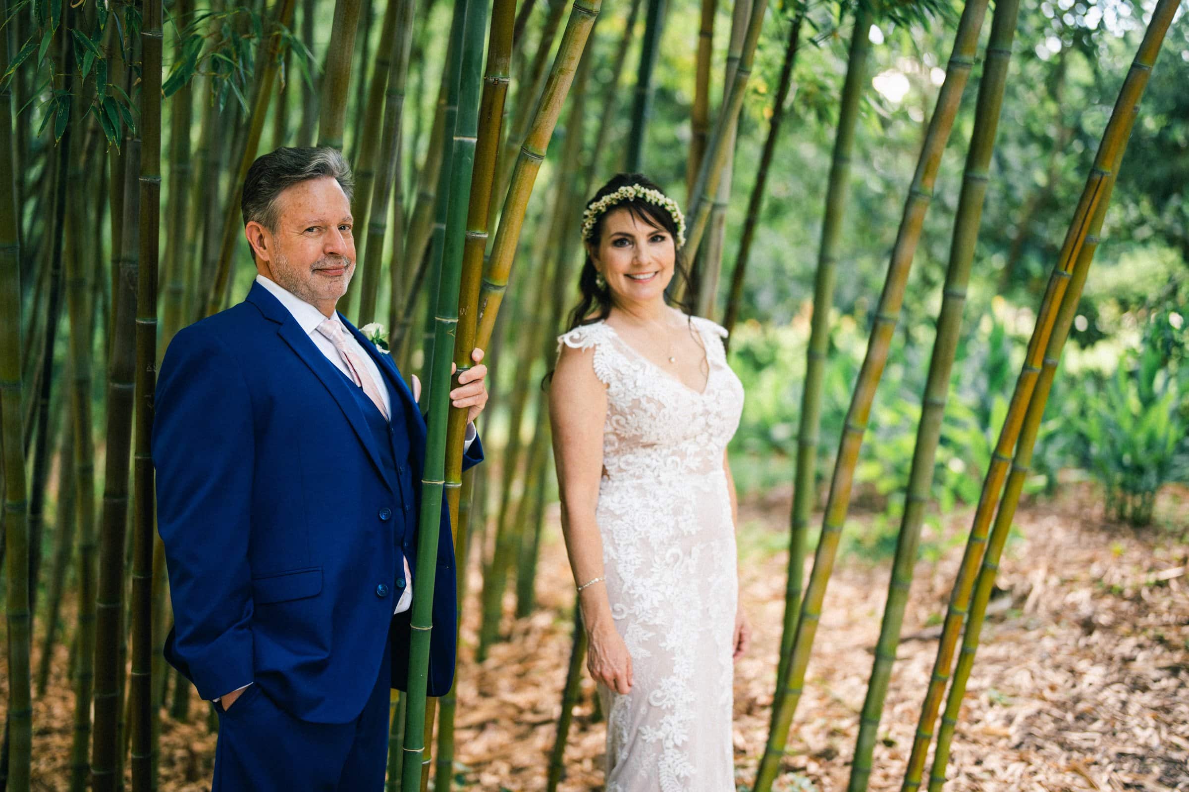 duke gardens wedding photography - ann & tim A man in a blue suit and a woman in a white lace dress stand amid tall bamboo stalks, embodying the essence of Duke Gardens wedding photography. The woman wears a leafy headpiece, and both appear happy, surrounded by lush greenery.