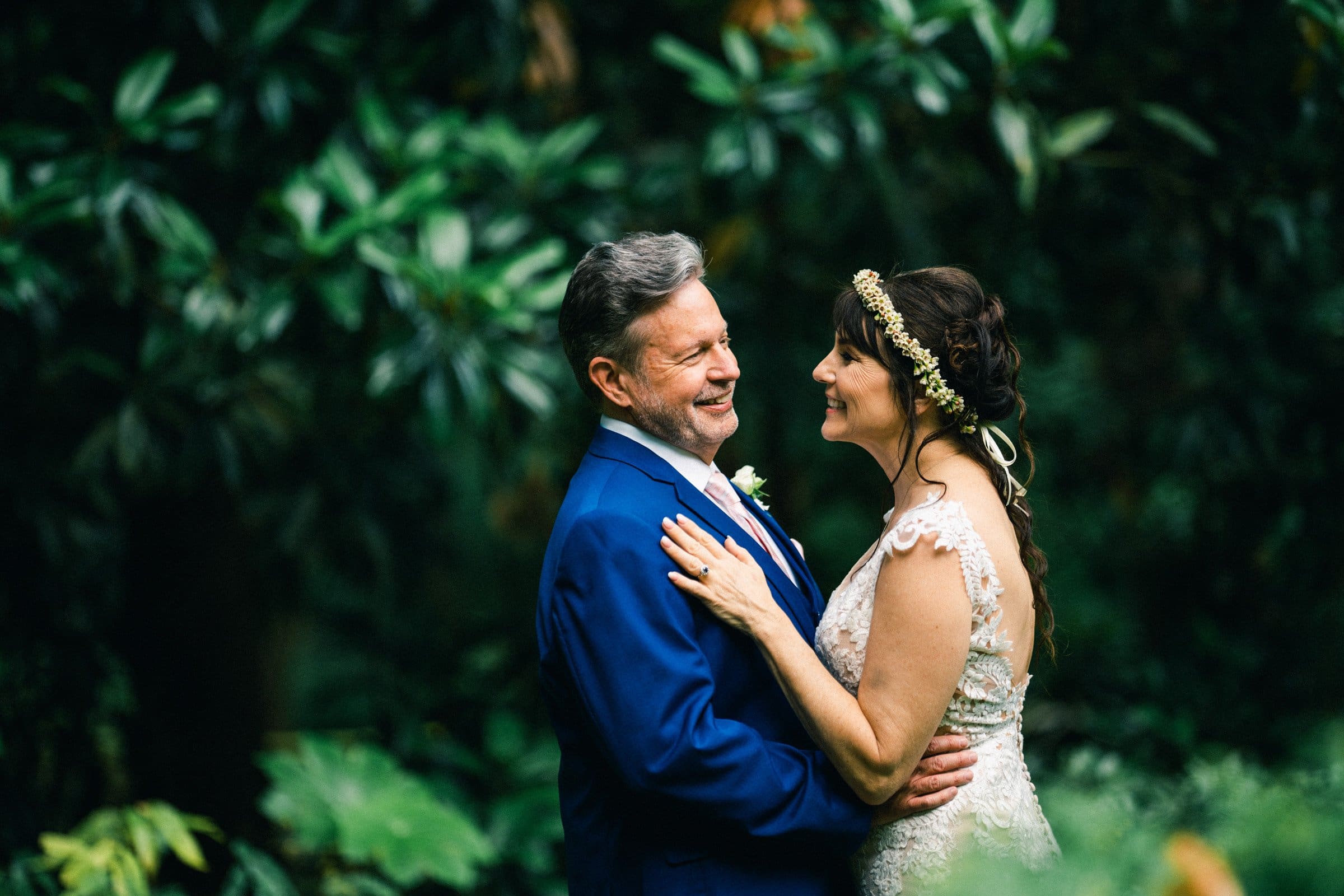 duke gardens wedding photography - ann & tim A couple in wedding attire stands smiling at each other amidst the lush greenery of Duke Gardens. The woman wears a lace dress and floral headpiece, while the man dons a blue suit with a white shirt, all beautifully captured in enchanting wedding photography surrounded by vibrant foliage.
