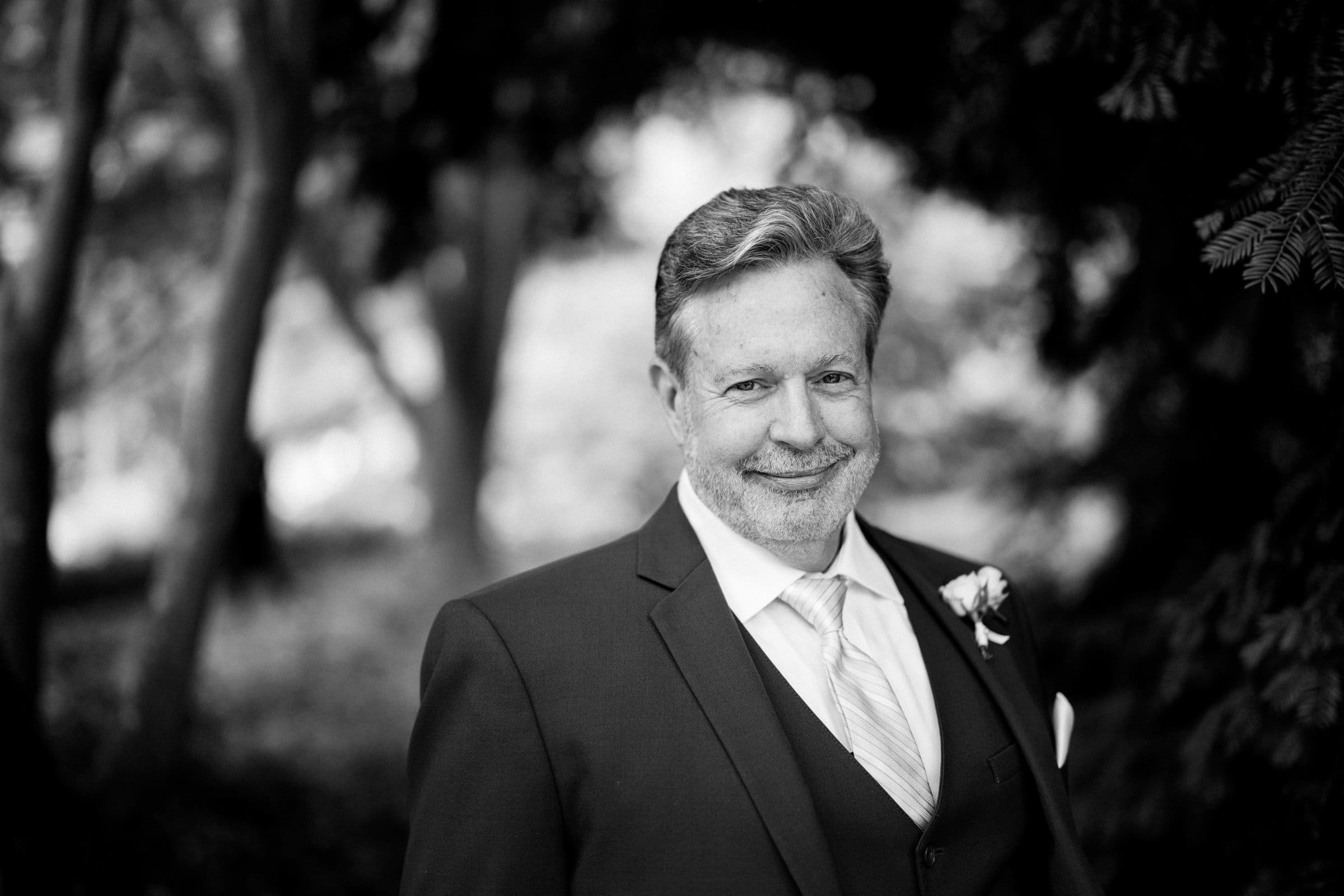 duke gardens wedding photography - ann & tim A man in a suit smiles at the camera, exuding charm with his short hair and beard. He wears a tie adorned with a boutonniere on his lapel. The blurred outdoor backdrop, suggestive of Duke Gardens, enhances the timeless elegance of this black-and-white wedding photography moment.