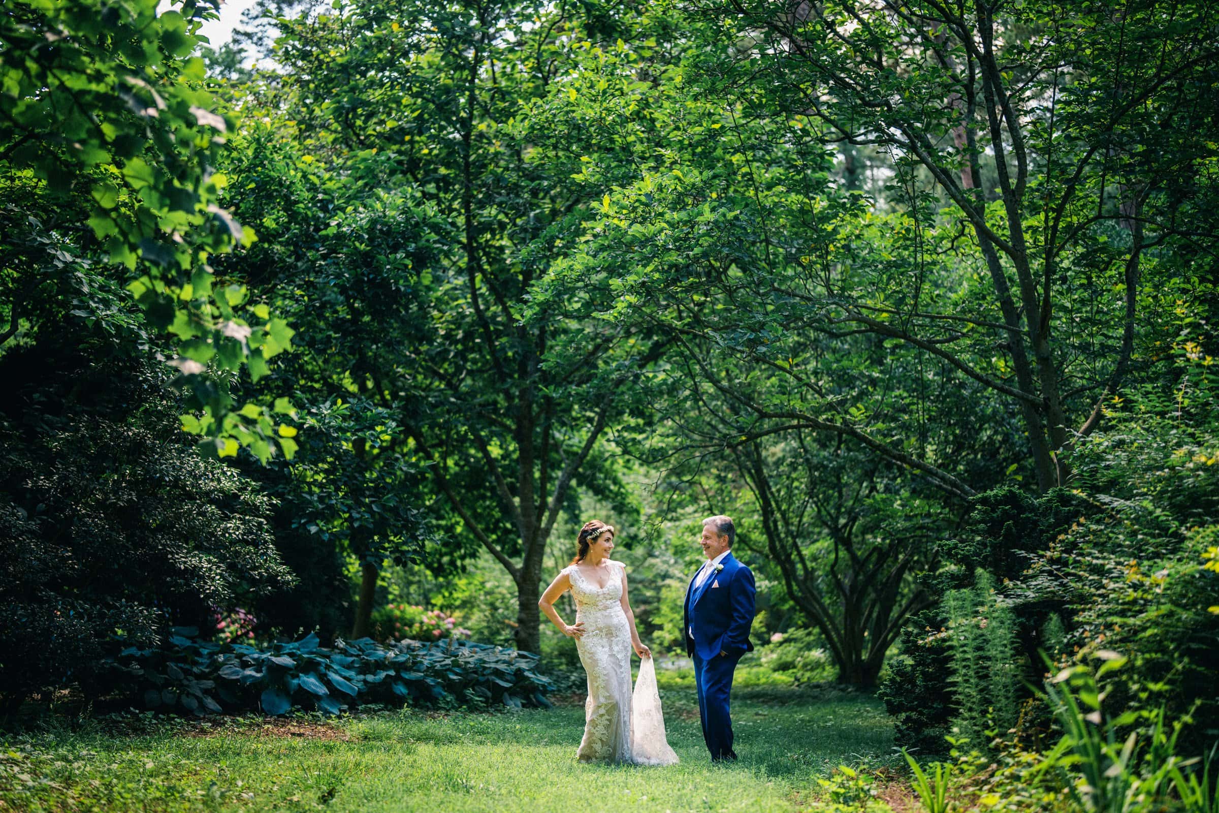 duke gardens wedding photography - ann & tim A bride and groom stand in a lush, green garden. The bride wears a white dress and holds its train, while the groom sports a blue suit. Sunlight filters through the trees at Duke Gardens, creating a serene, romantic atmosphere perfect for wedding photography.