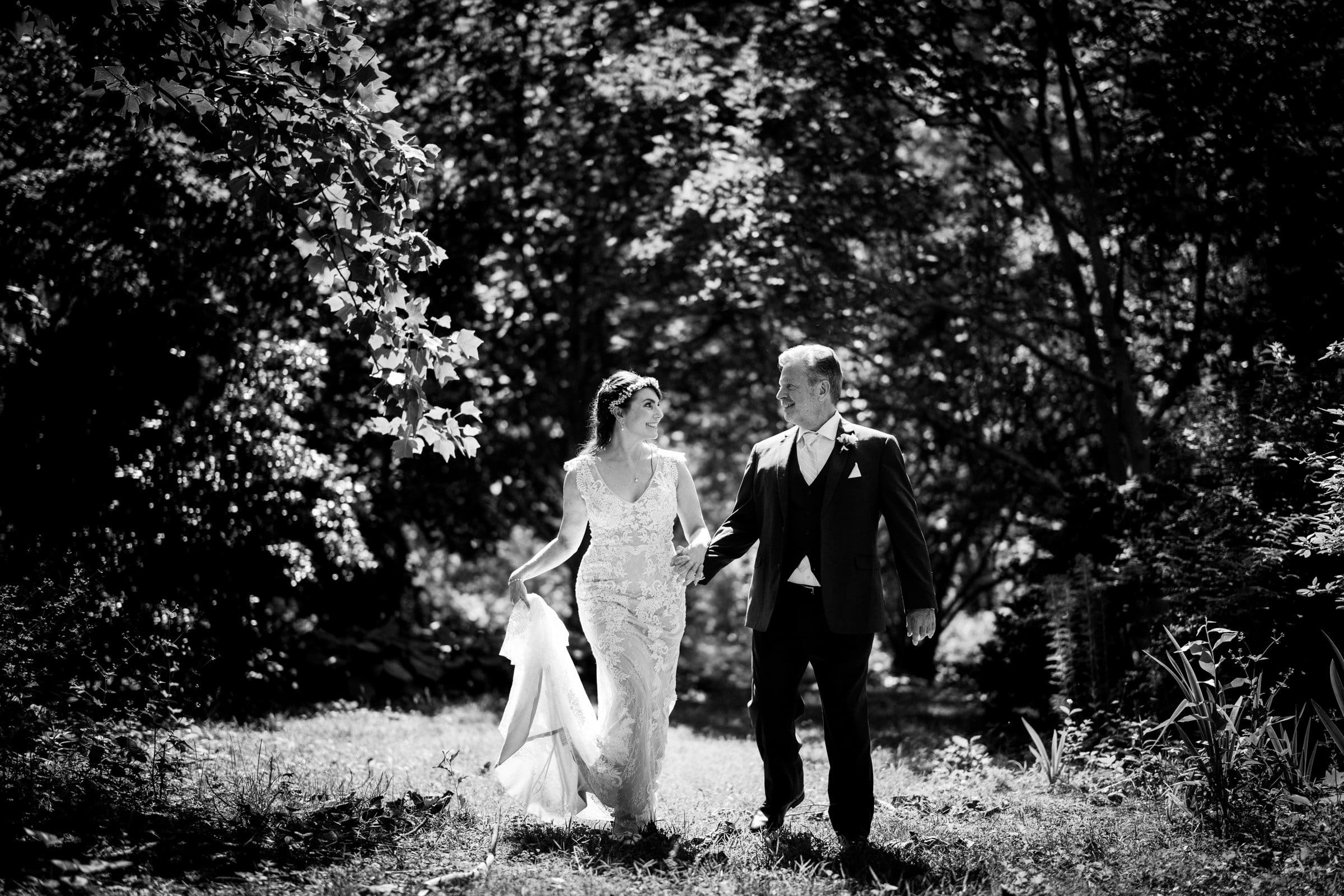 A black and white photo captures a bride and groom in Duke Gardens, walking through a wooded area. The bride, holding her train, wears an elegant dress, while the groom complements her in a suit. They gaze at each other, enveloped by sunlight filtering through the trees.
