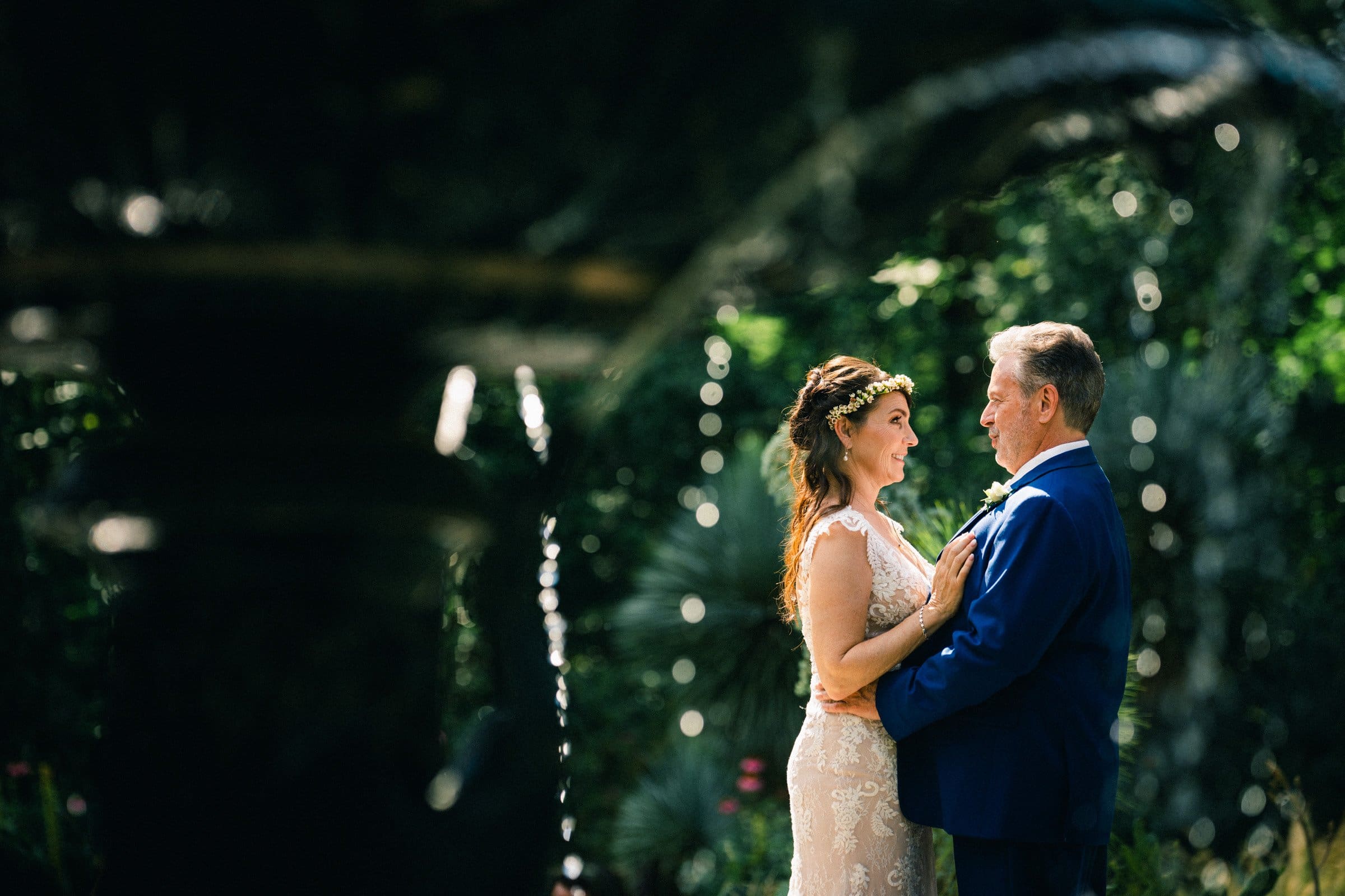duke gardens wedding photography - ann & tim A couple in formal attire stands facing each other in Duke Gardens, capturing the essence of wedding photography. The woman, with a floral headpiece and lace dress, gazes at the man in a blue suit. Dappled sunlight and a blurred fountain enhance this romantic scene.