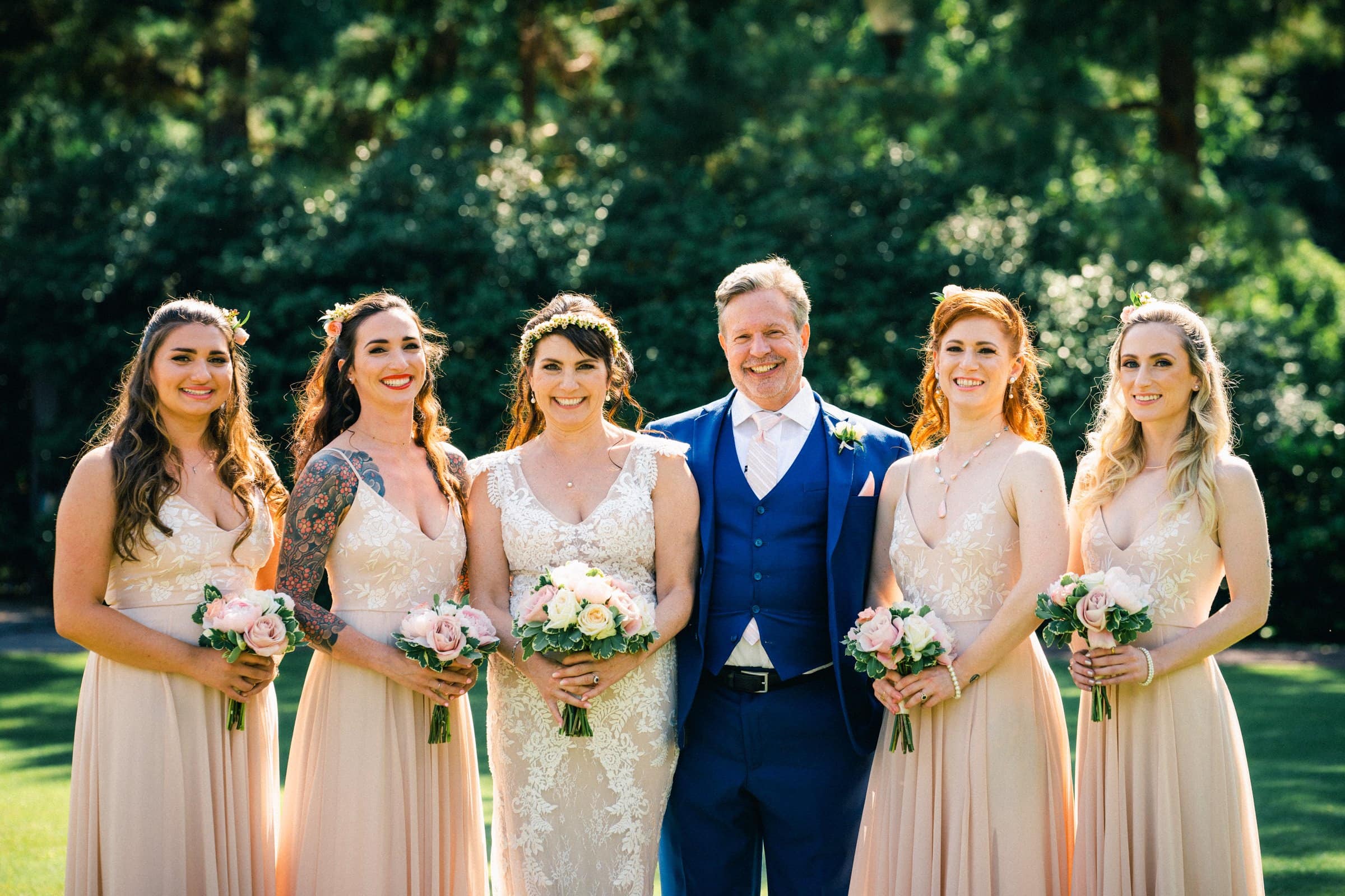 duke gardens wedding photography - ann & tim A wedding party poses outside in Duke Gardens. The bride, in a lace gown, and groom, in a blue suit, stand together holding bouquets. They are surrounded by five women in peach dresses, each clutching a bouquet amidst the lush greenery that forms the perfect backdrop for their magical day.