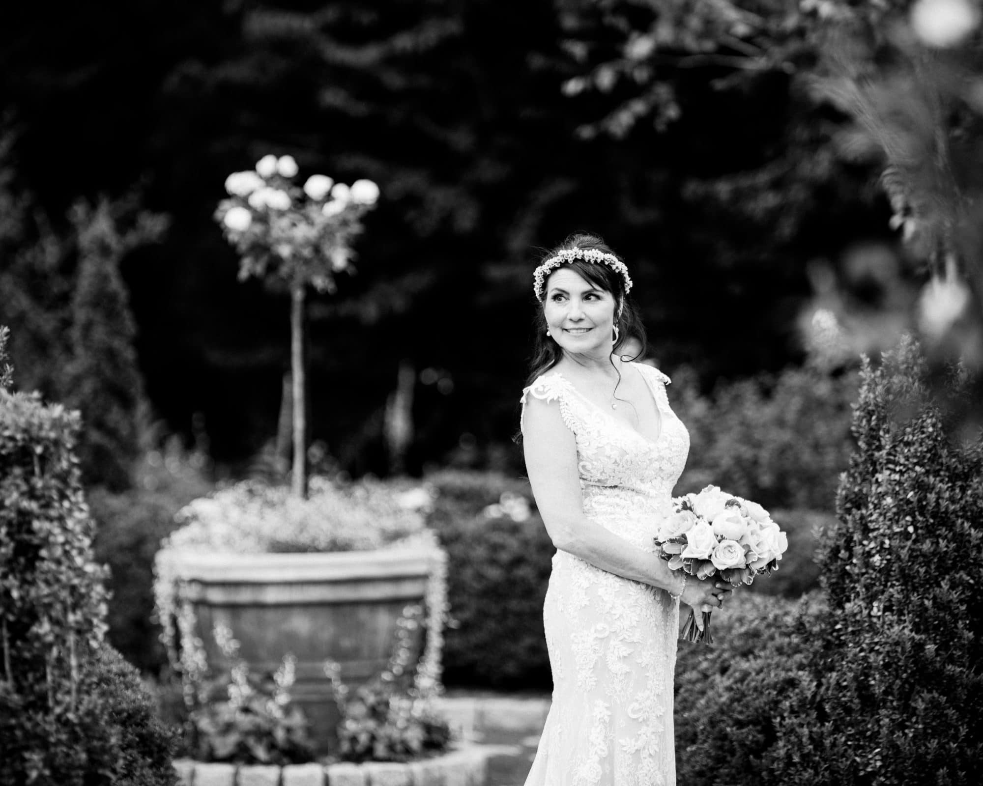 duke gardens wedding photography - ann & tim A bride in a white lace gown and floral headpiece graces the garden of Duke Gardens, holding a bouquet. She gazes to the side with a soft smile amid lush greenery and a large potted plant. The image, captured in black and white, embodies timeless wedding photography.
